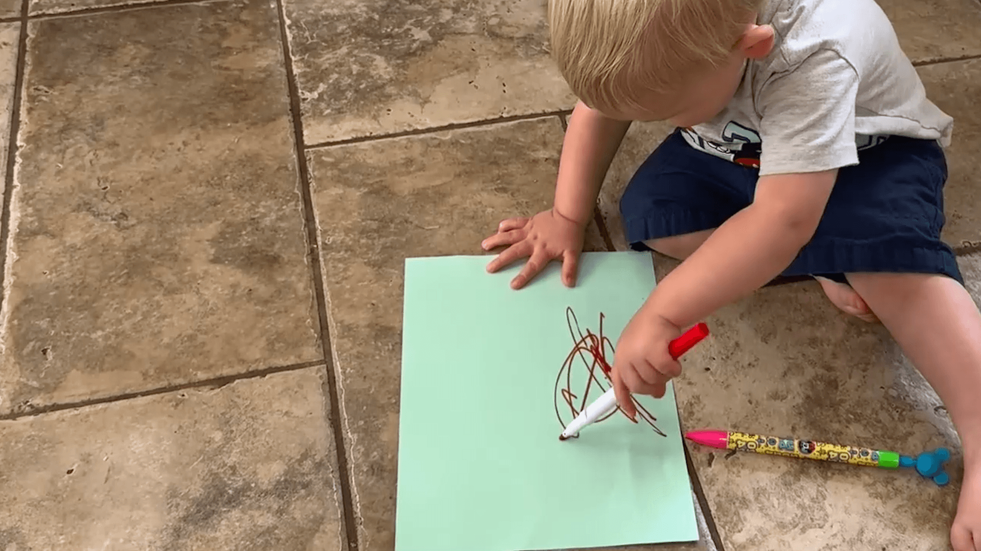 18-24-month-old toddler boy uses the digital pronate grasp to scribble on a sheet of green paper while sitting on the floor.