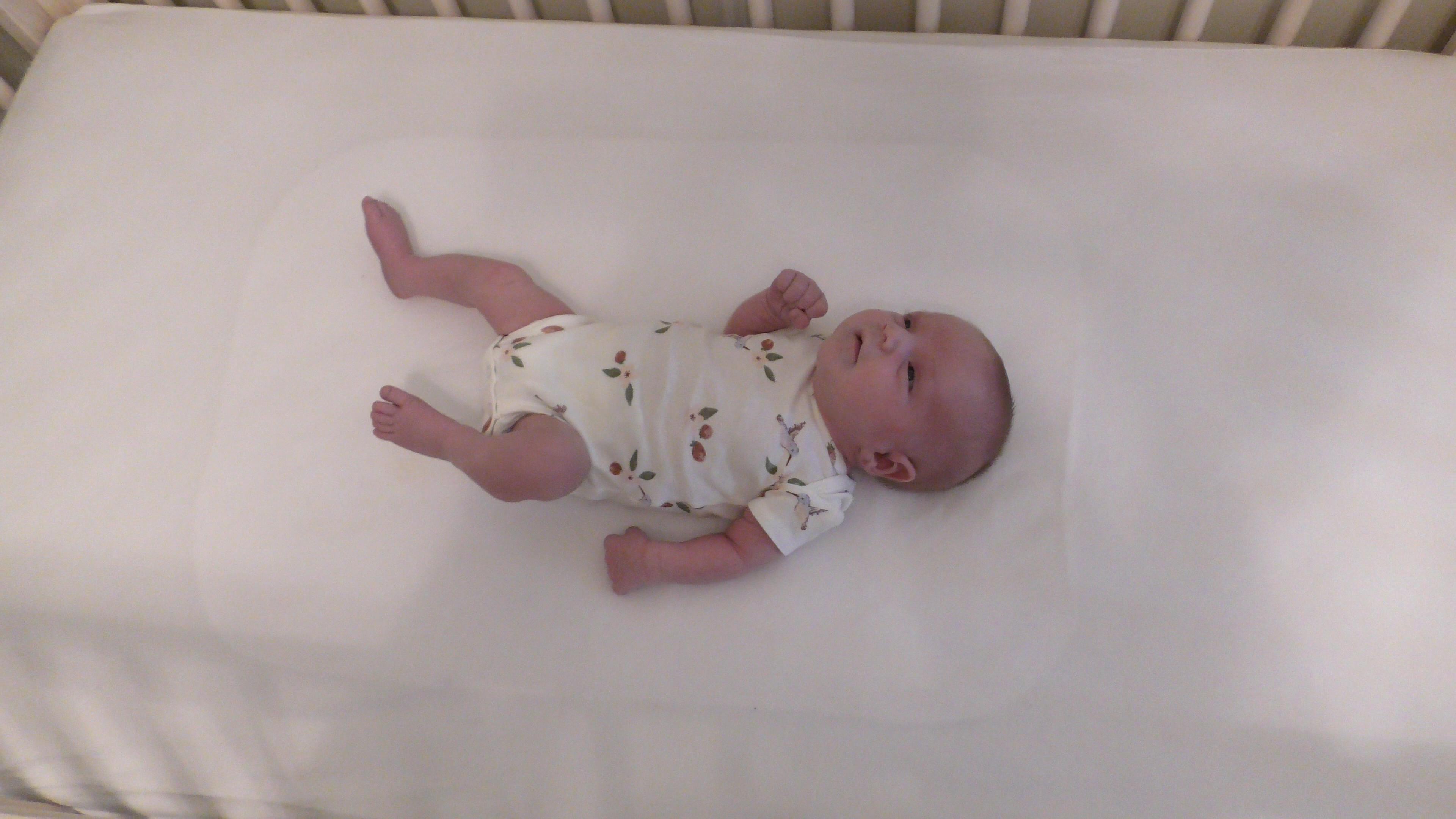Tiny newborn on a white crib mattress looking upward with sleepy eyes.