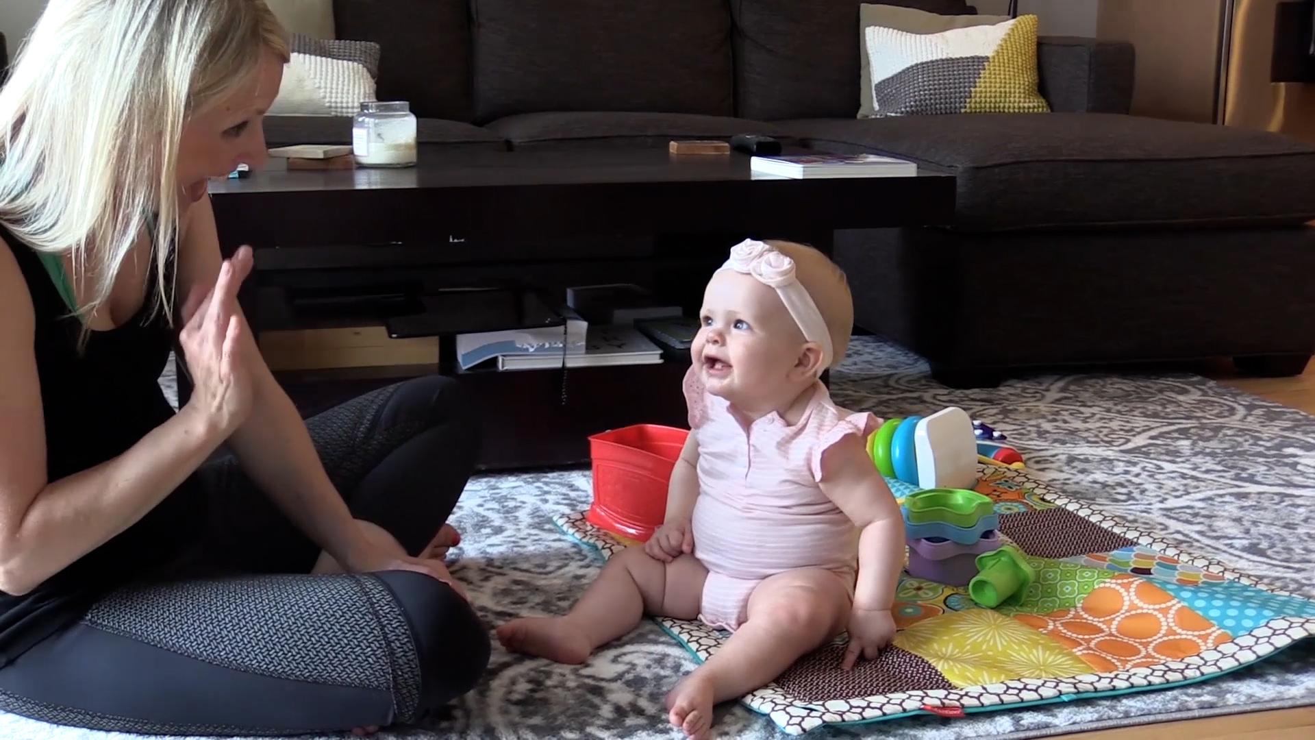  Mom waving at talking baby girl as they sit on living room floor with a baby playmat and toys.