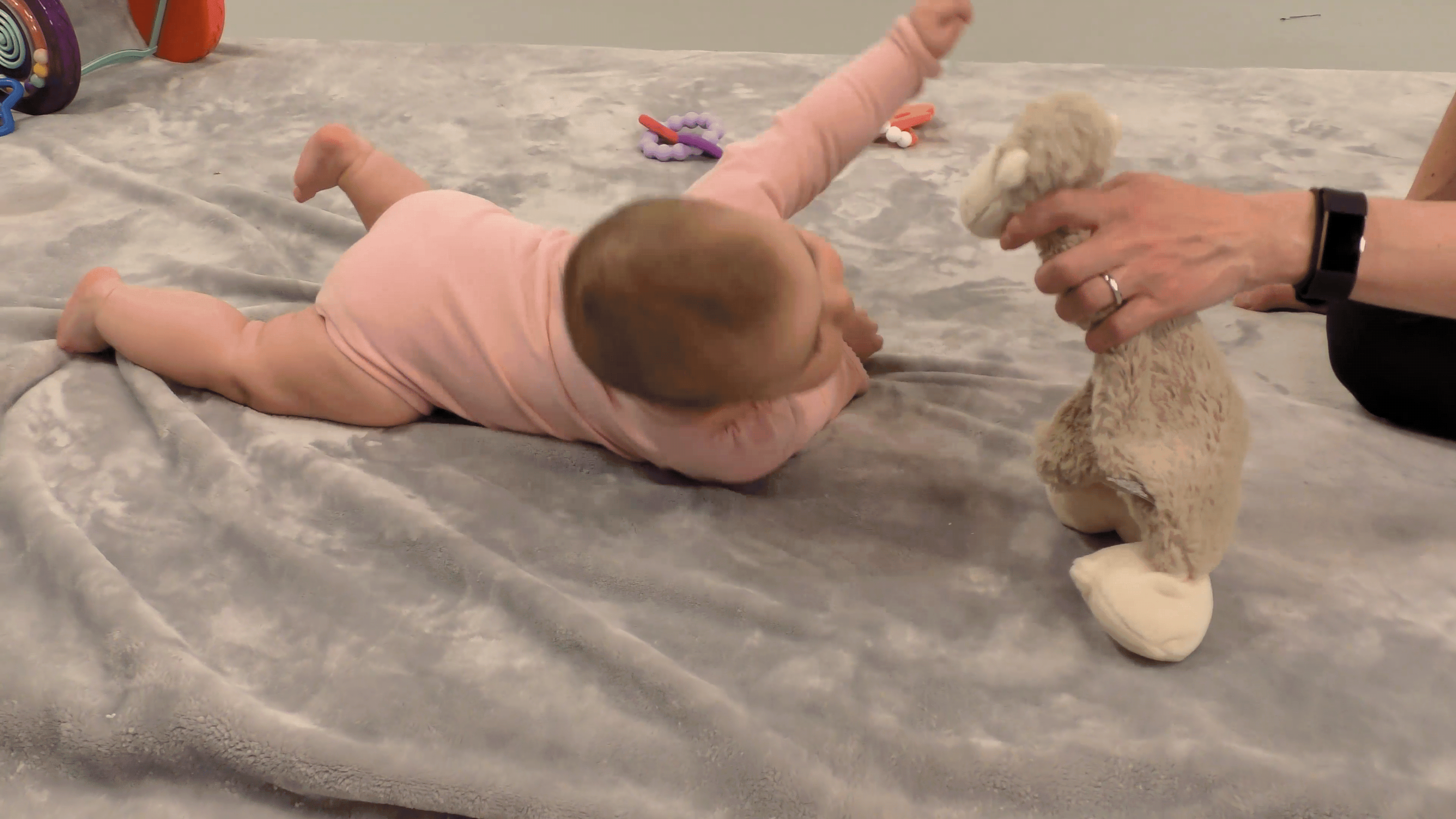 Baby girl in pink onesie attempting to roll over while a caregiver shows her a stuffed animal.