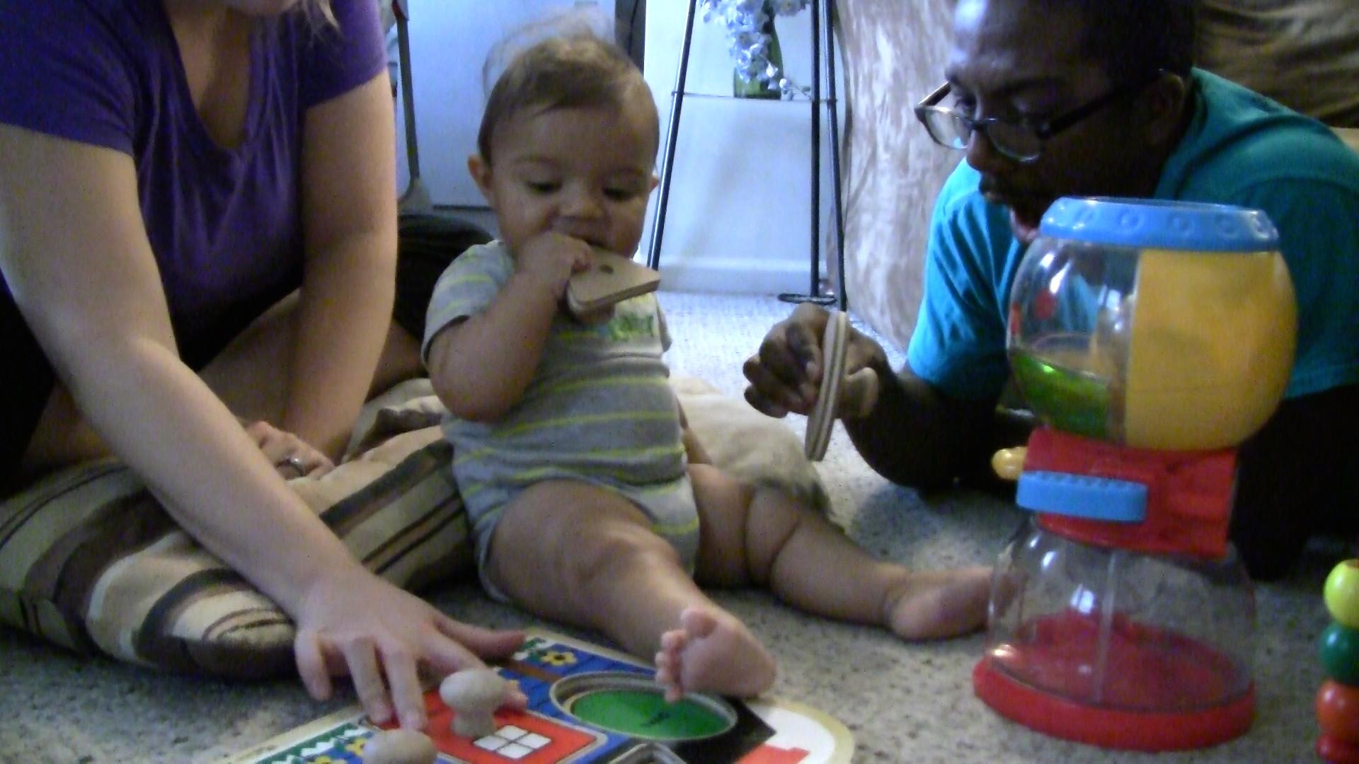 A baby boy sits on the floor between his parents while holding a triangle puzzle piece in his mouth. A baby shape puzzle and other toys surround him.