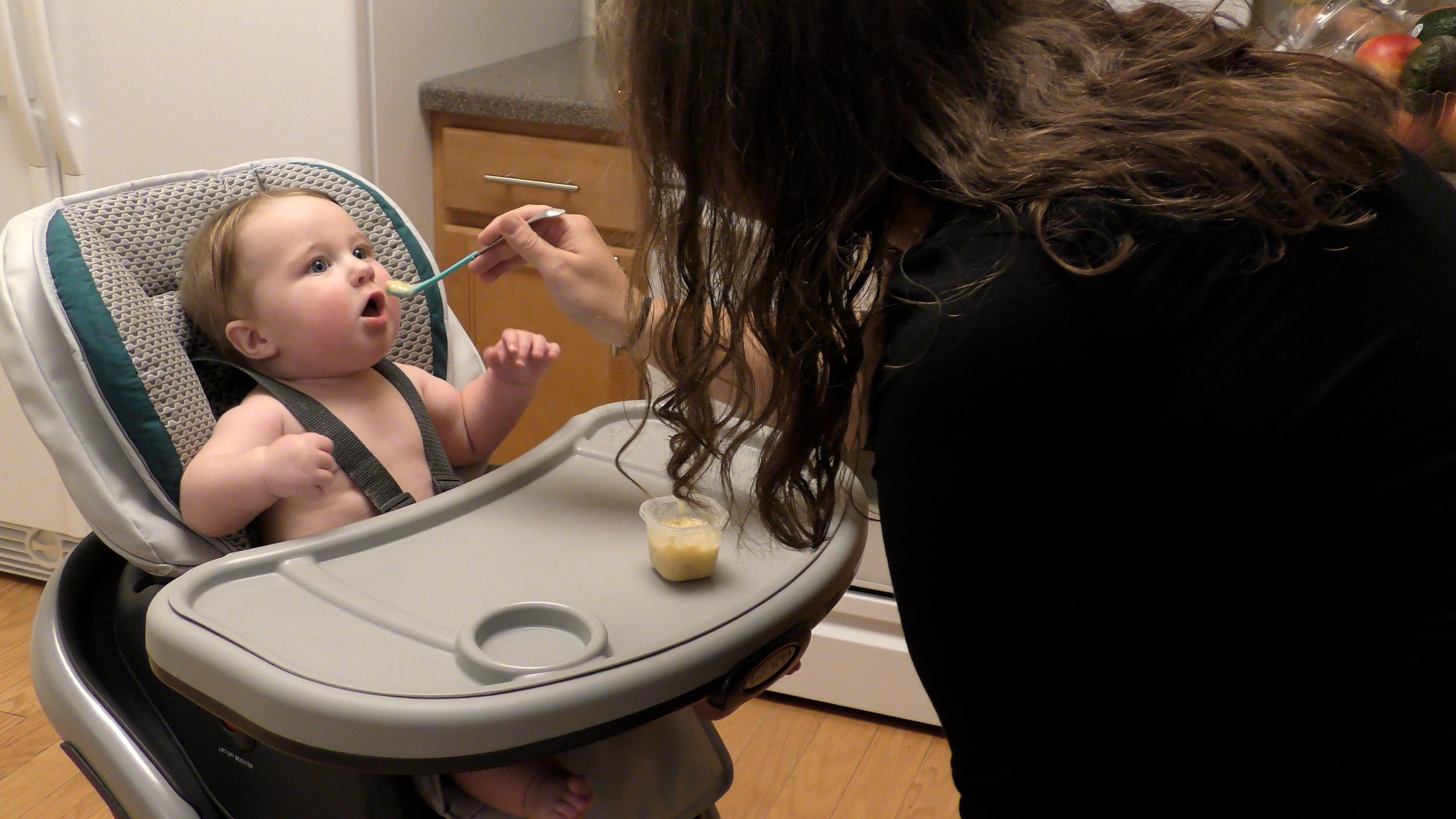 Mom spoon feeding baby boy mashed banana while he sits in his high chair.