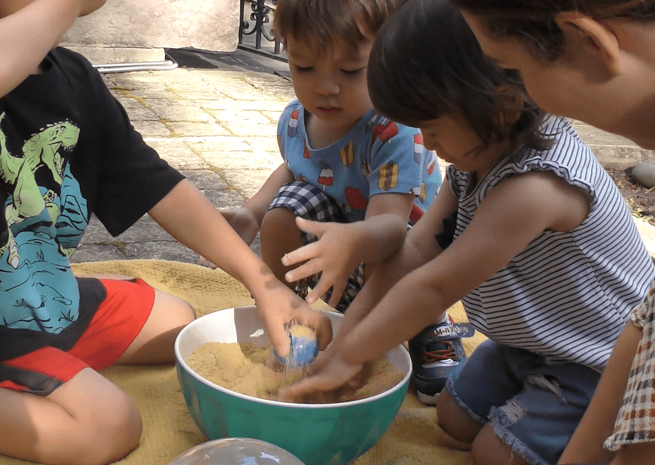 Two toddlers and a preschooler sitting around a bowl of crushed cereal using their hands to search for items buried in it.