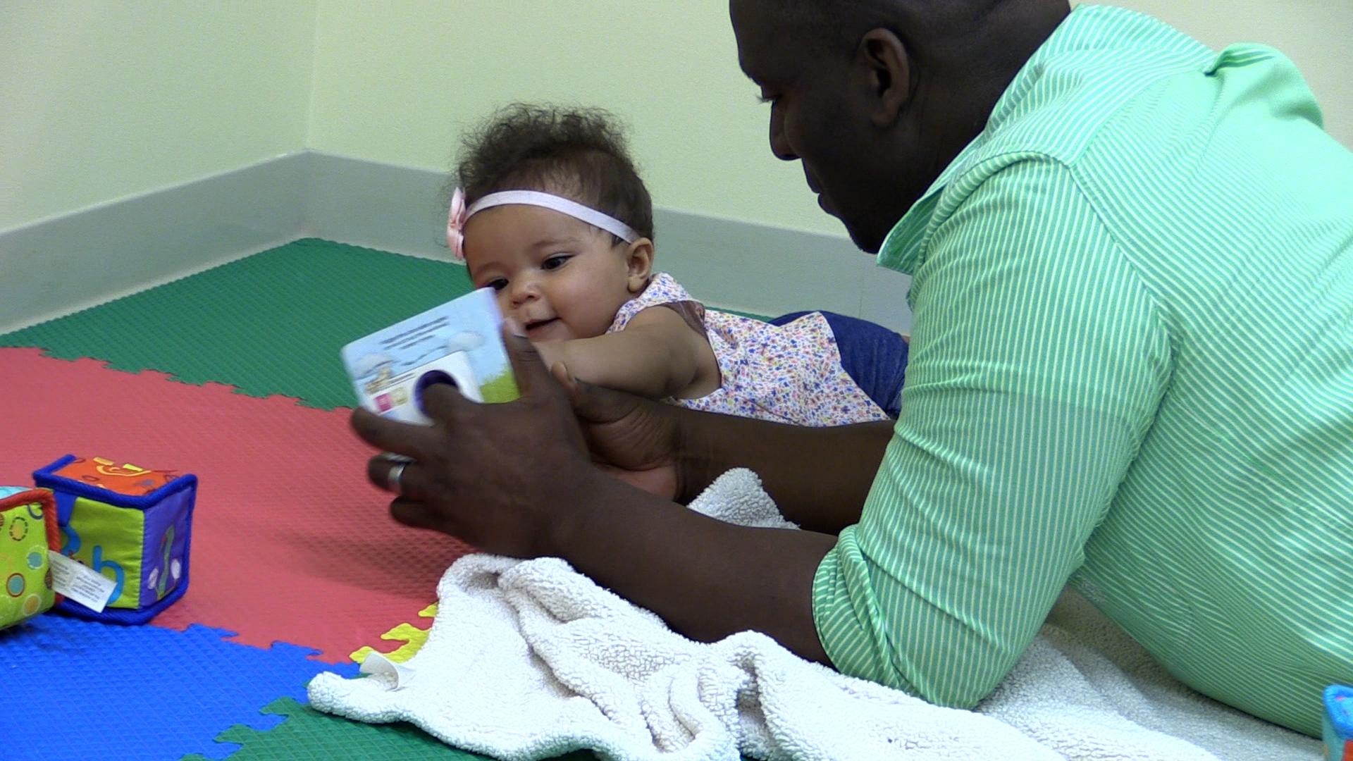 Dad reading to baby girl while both lie on their tummies on top of a multi-colored puzzle foam mat. 
