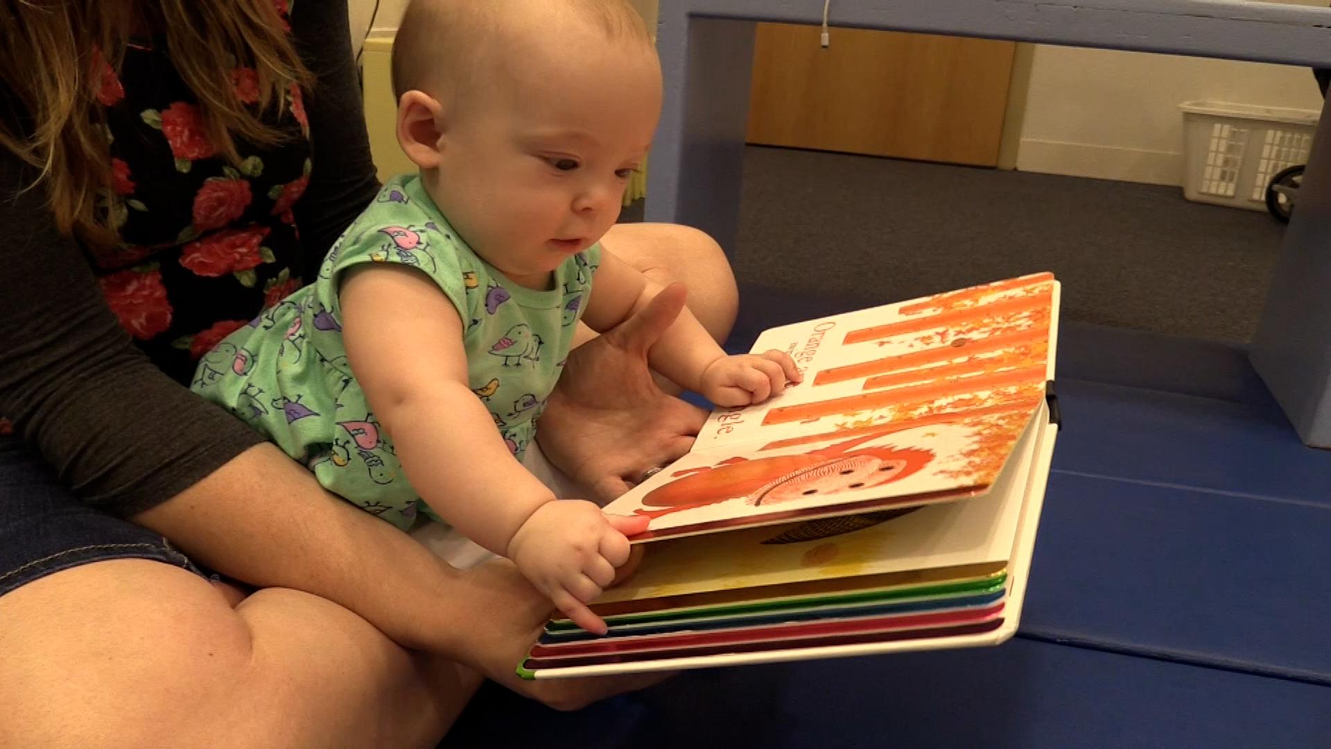 Baby girl in a mint green, bird-patterned dress leaning over and holding onto the pages of a baby board book. She is sitting in a woman’s lap on the floor.
