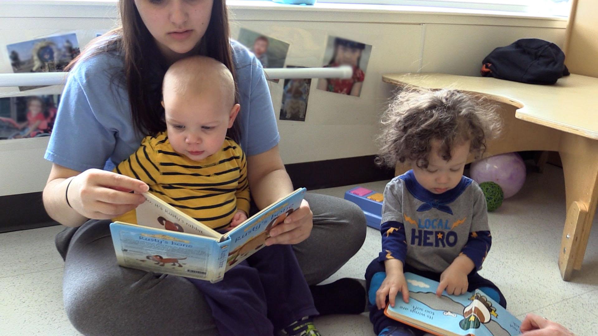 A toddler boy in a yellow-and-black striped shirt sits in a woman’s lap as she reads a book to him on the floor. Another toddler boy sits by himself to the right of them, pointing to a picture in a book he is reading with a person out of frame.