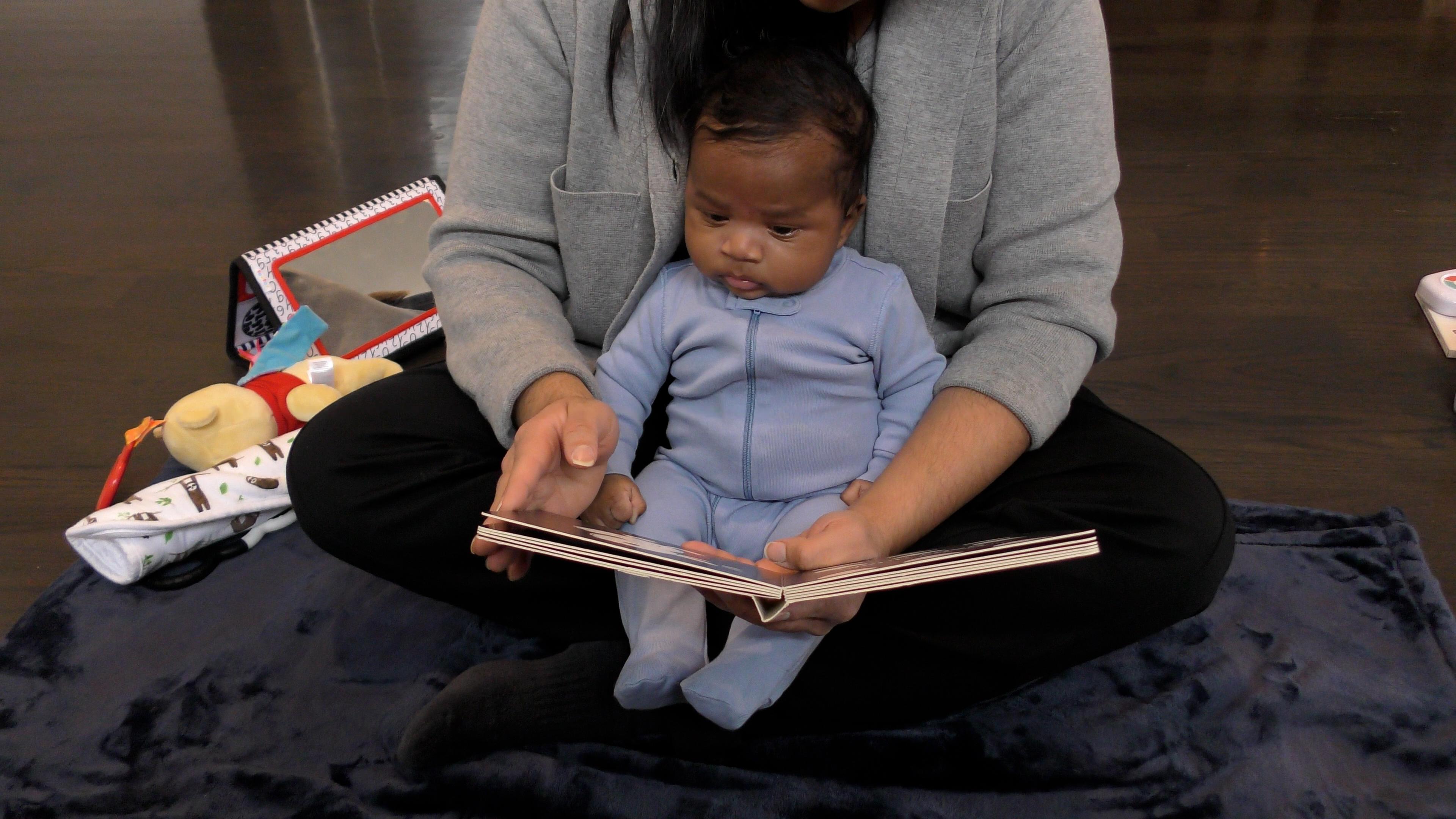 Newborn baby boy in blue onesie sitting up in a woman’s criss-crossed lap, looking down at the board book she’s reading to him.