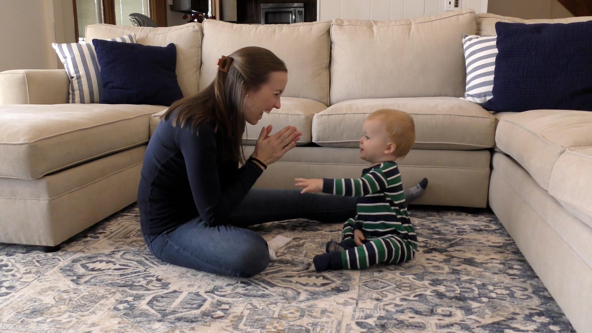 Smiling mom shows baby boy how to clap as they sit across from each other on a living room floor surrounded by a sectional couch.