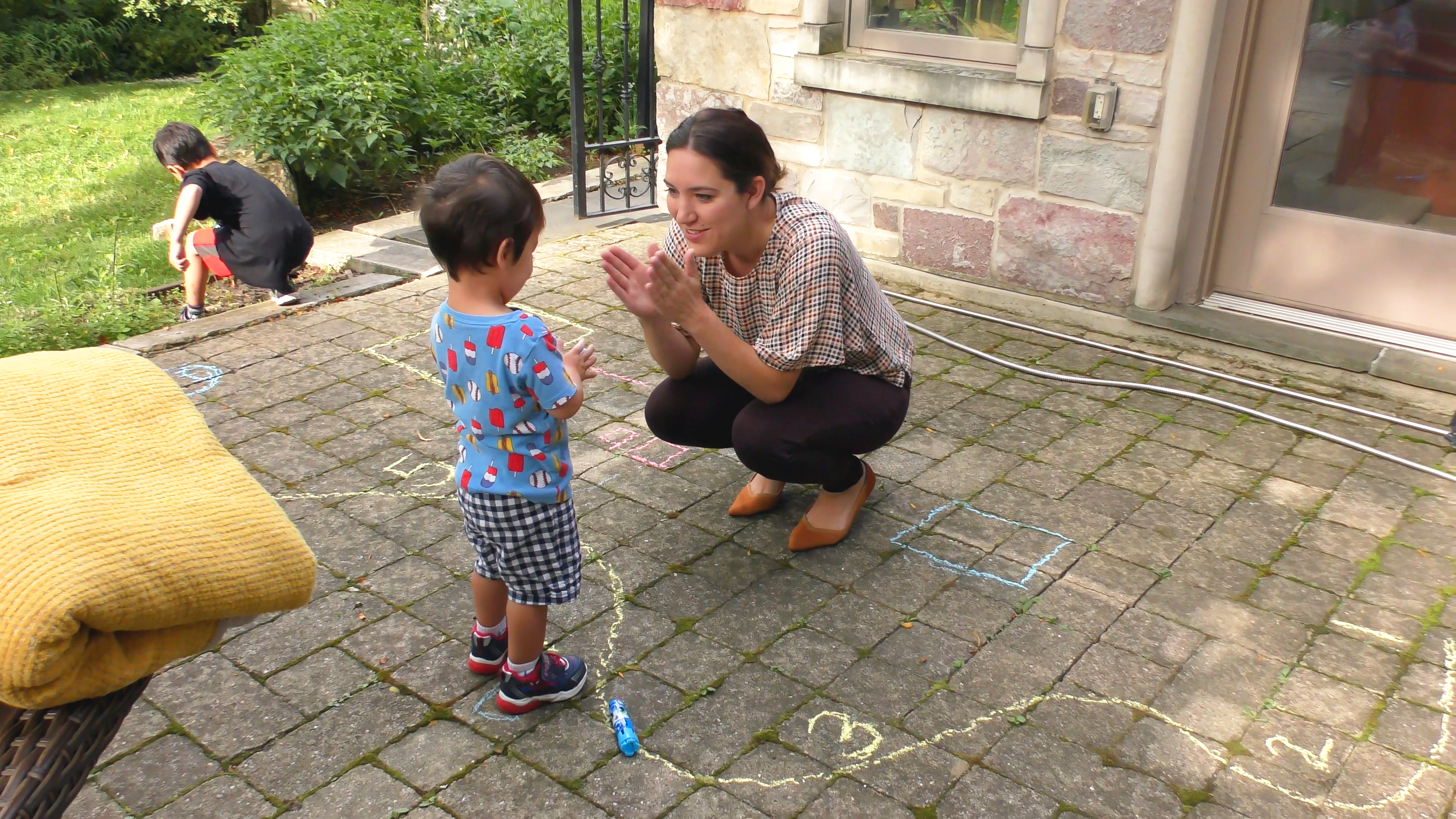Mom squatting in front of her toddler son and encouraging him with clapping. They stand on a patio with chalk drawings on it.