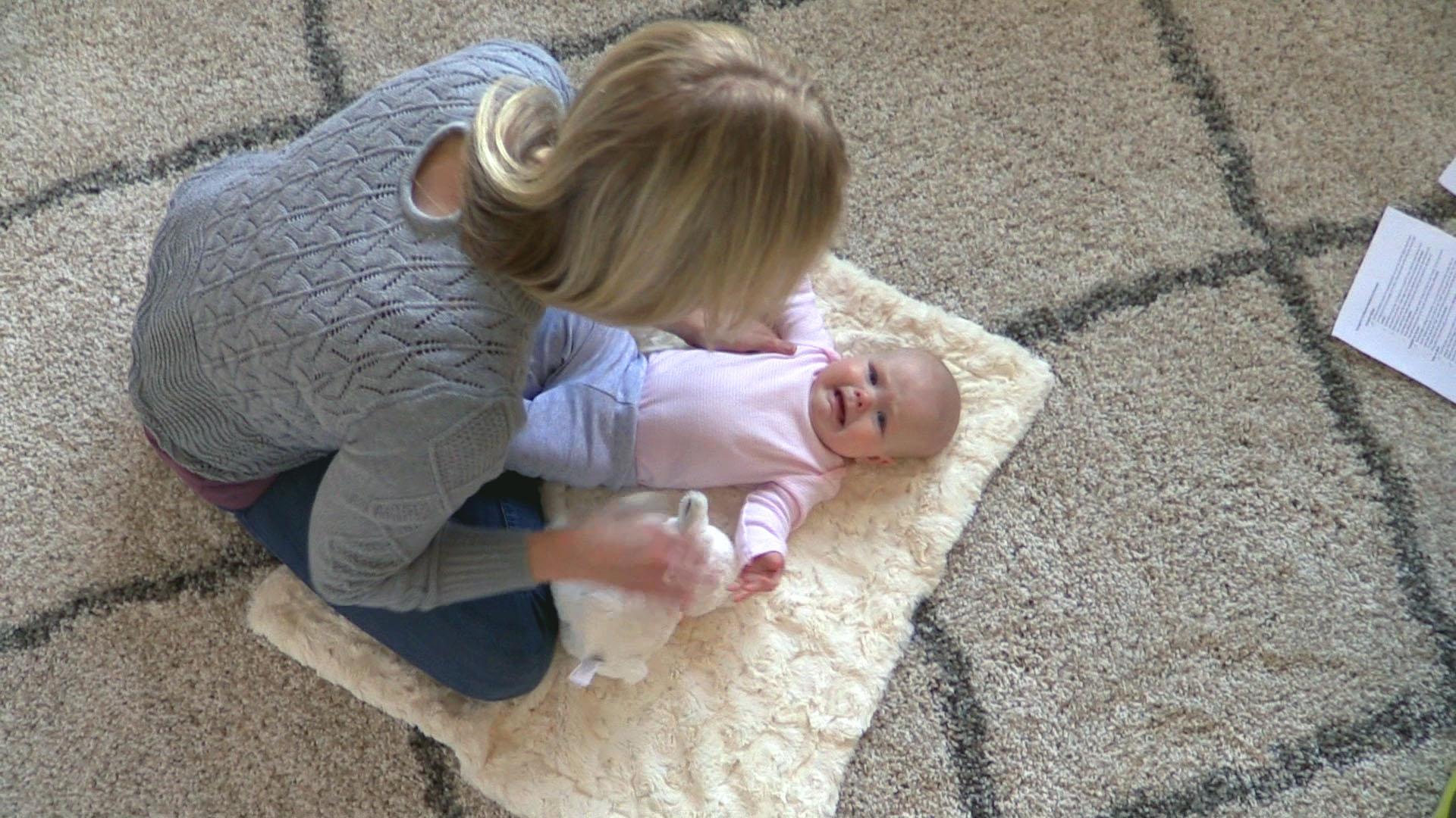 Top-down view of a blond-haired mother sitting cross-legged on the floor and leaning over her crying baby girl, who is lying on her back atop a cream-colored blanket.