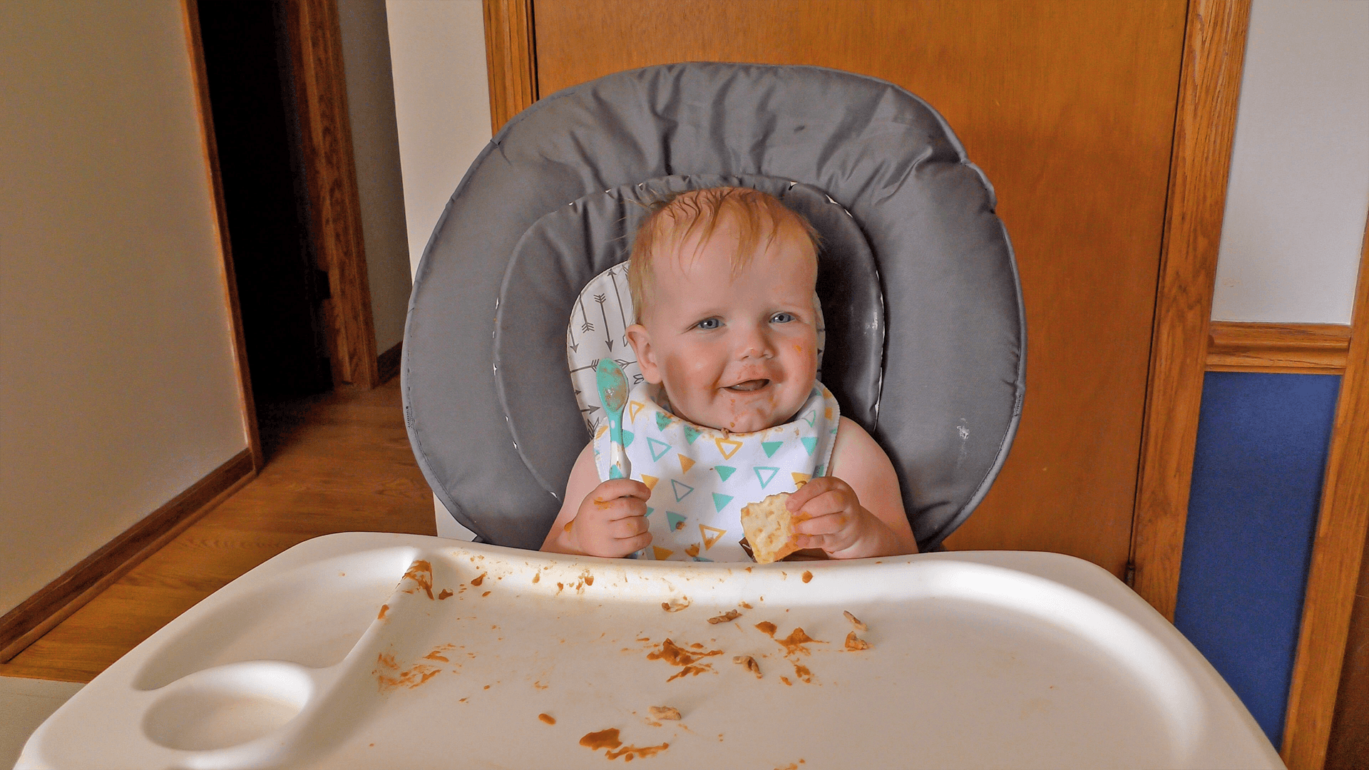6-9-month-old baby boy smiling ahead while holding a spoon and piece of bread in front of a messy high chair tray.