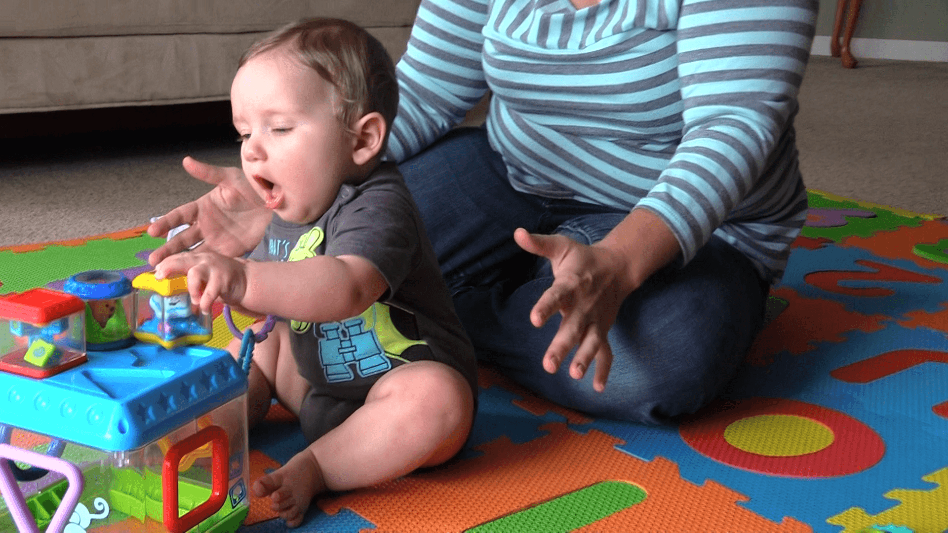 4-6-month-old baby boy grabbing a star-shaped baby toy with his whole palm. He sits on a colorful puzzle-piece playmat in front of his mother.