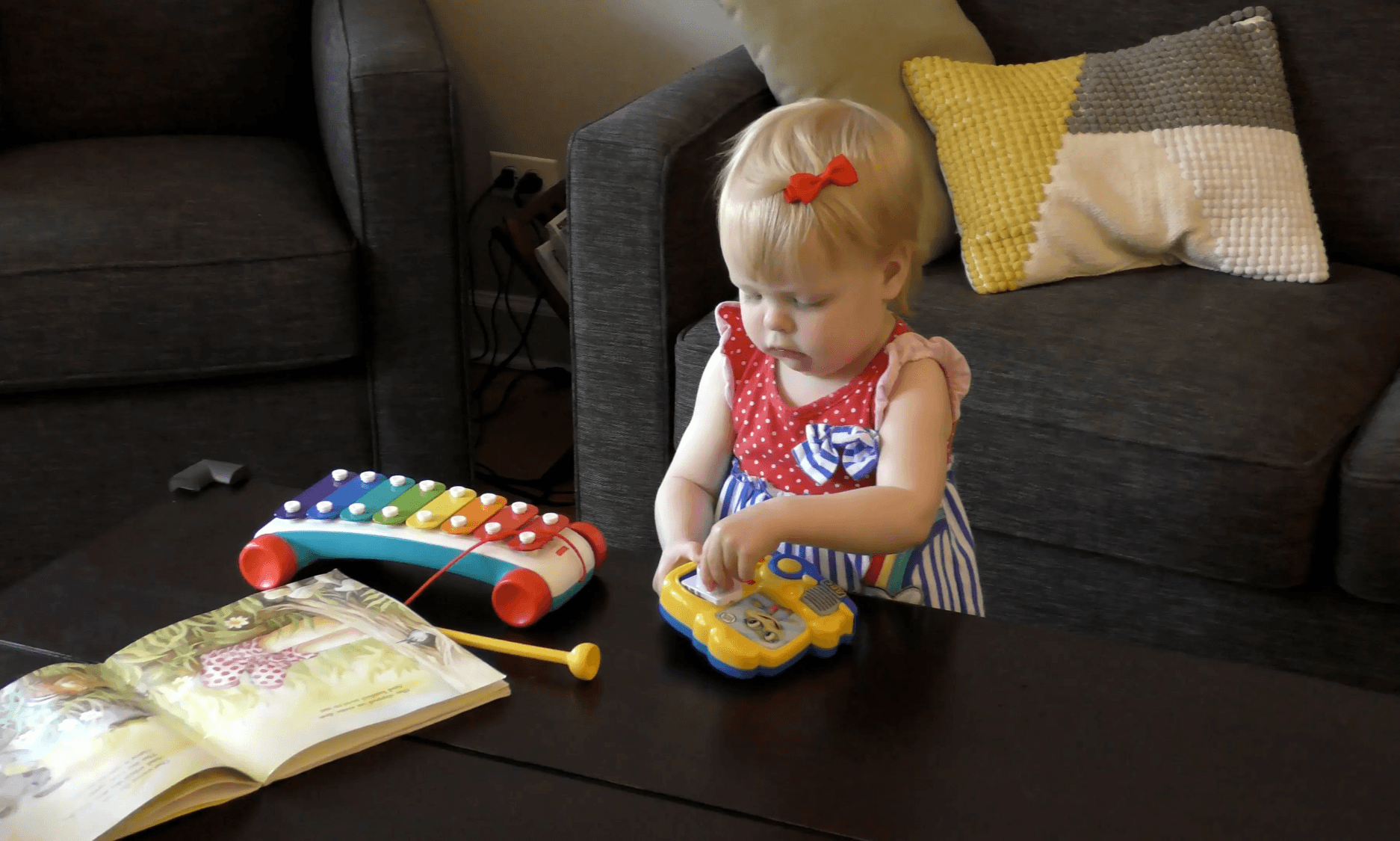 Toddler girl playing independently with sound-making toys and a book.