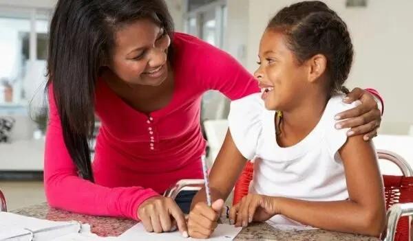 A smiling woman in a red shirt helps a young girl with schoolwork. The girl holds a pencil and smiles during their conversation.