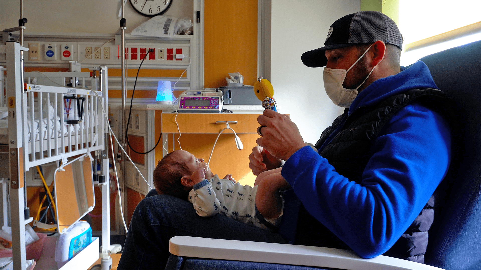 Dad waving soft toy at preterm baby boy lying on his lap. They are sitting in a NICU room surrounded by a crib and medical equipment.