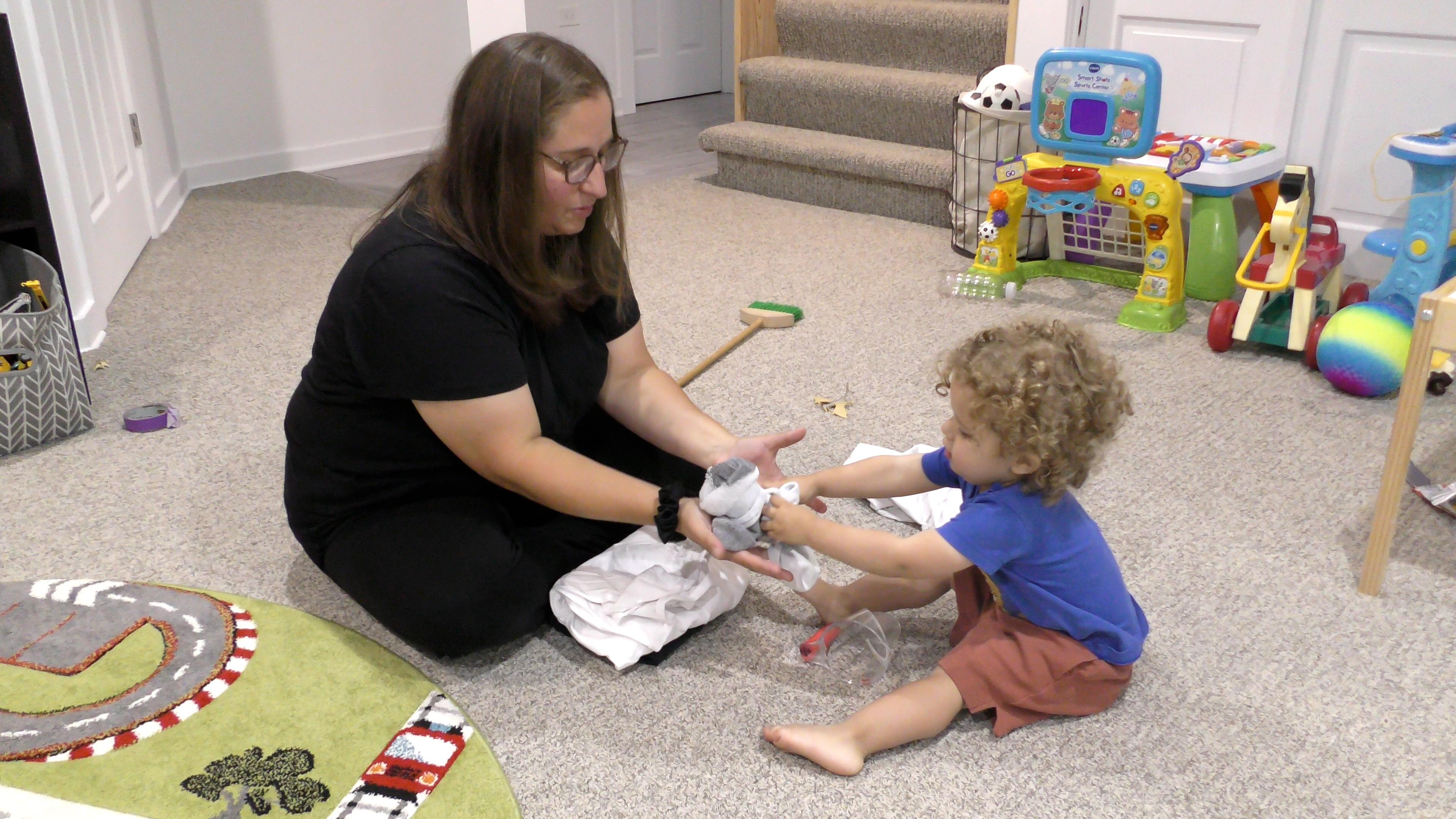 Toddler boy handing socks to his mom as she folds laundry on the playroom floor.