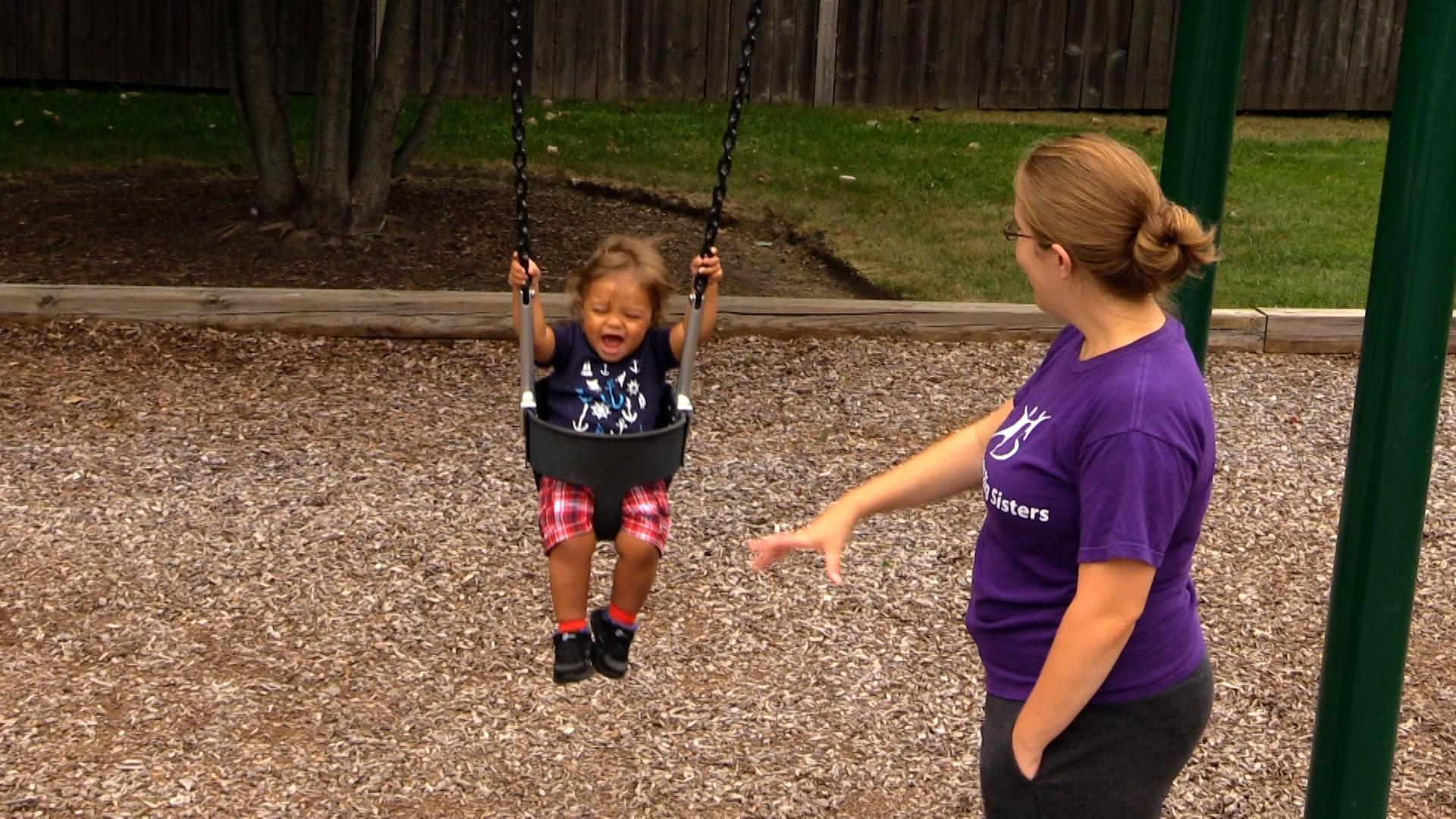 A mom in a purple shirt pushes her toddler son in a swing. The boy has an expression of excitement on his face as he holds onto the swing chains.