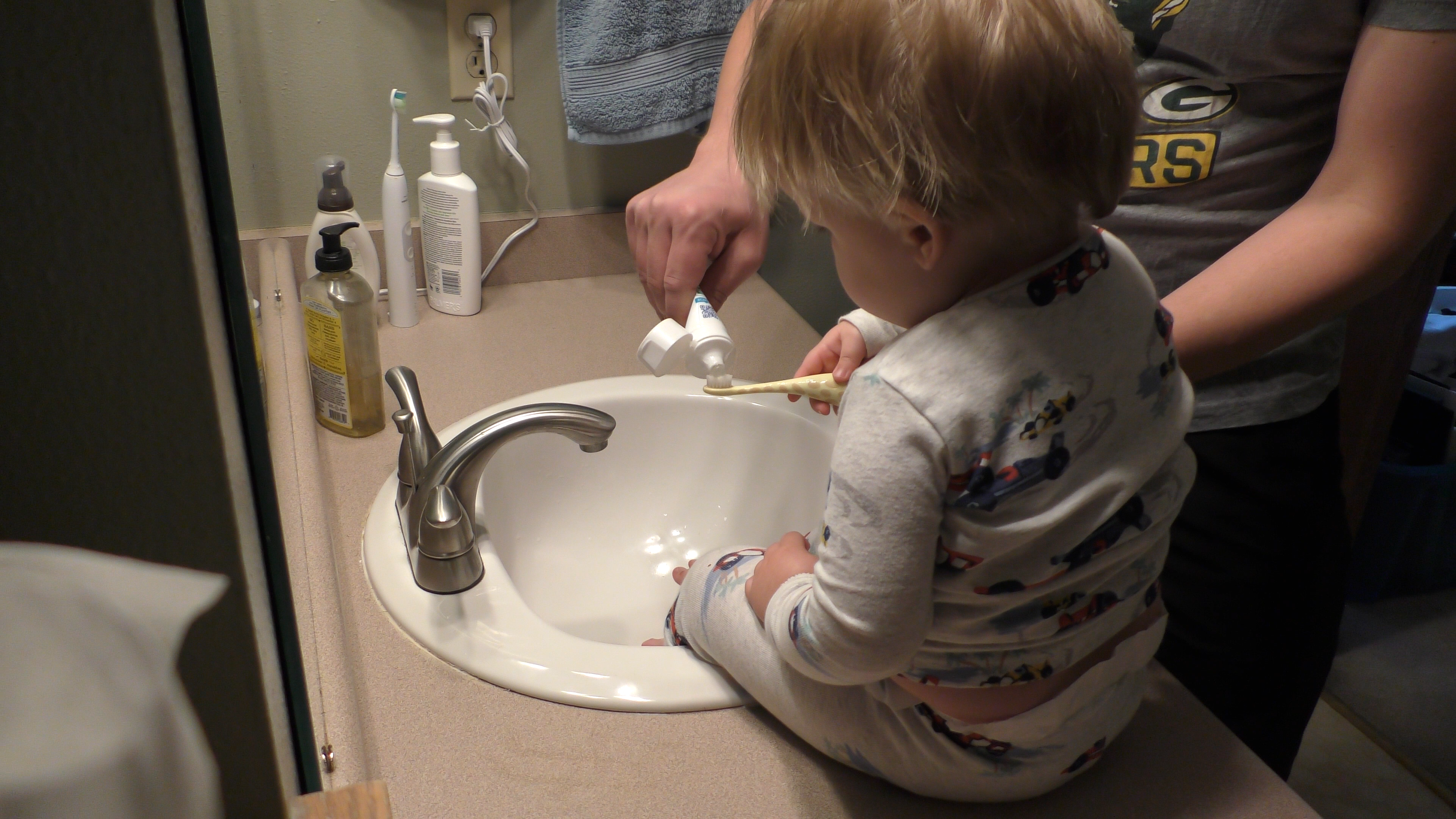 Parent helping toddler put the right amount of toothpaste on his brush before brushing his teeth.