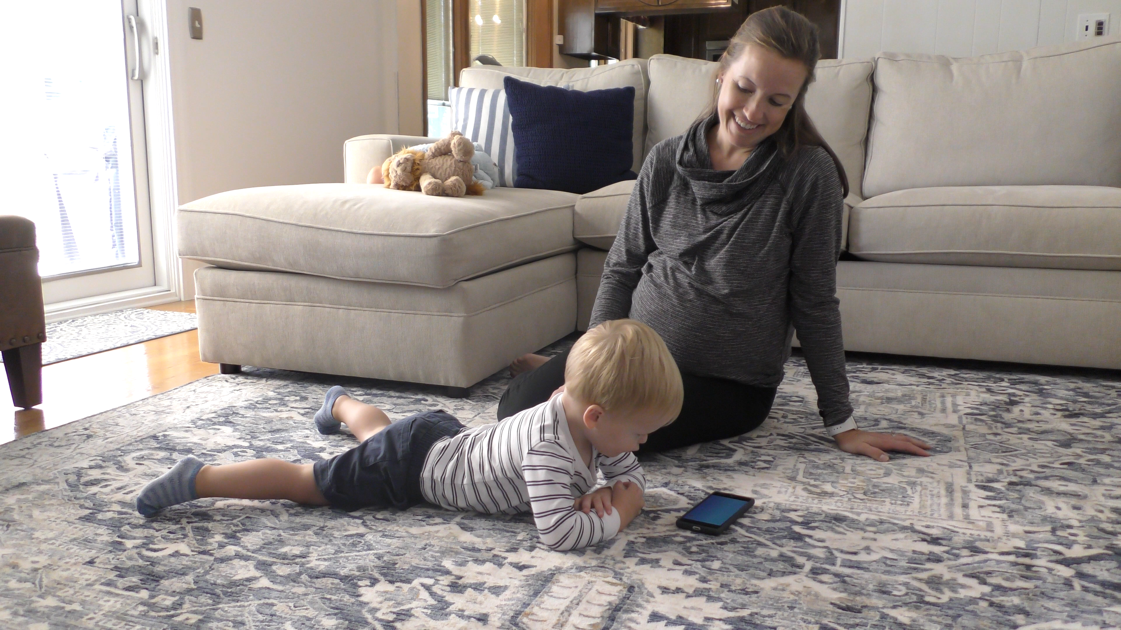 A mom and toddler sit on a living room floor and watch a video on a phone together.