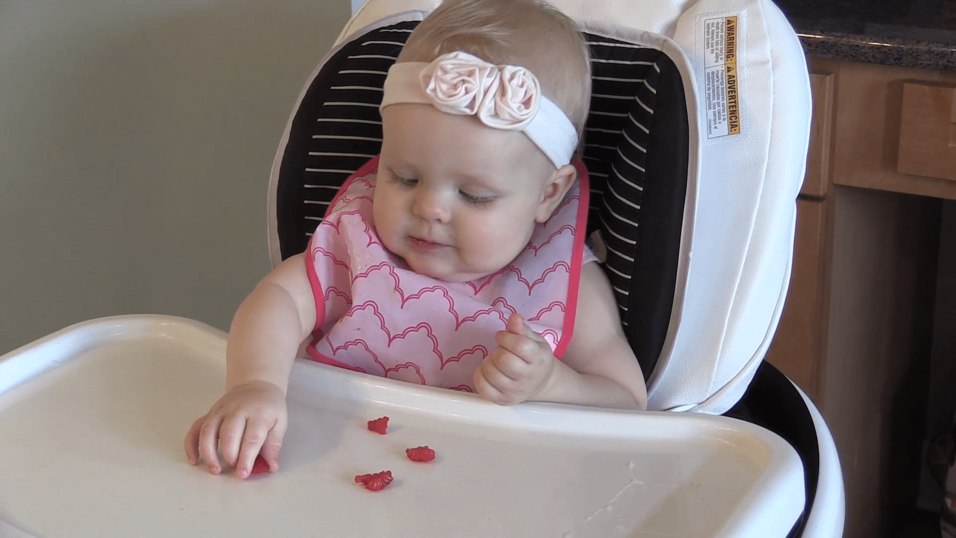 Baby girl in light pink rose headband and pink bib sitting in high chair using a crude pincer grasp to pick up a piece of strawberry.