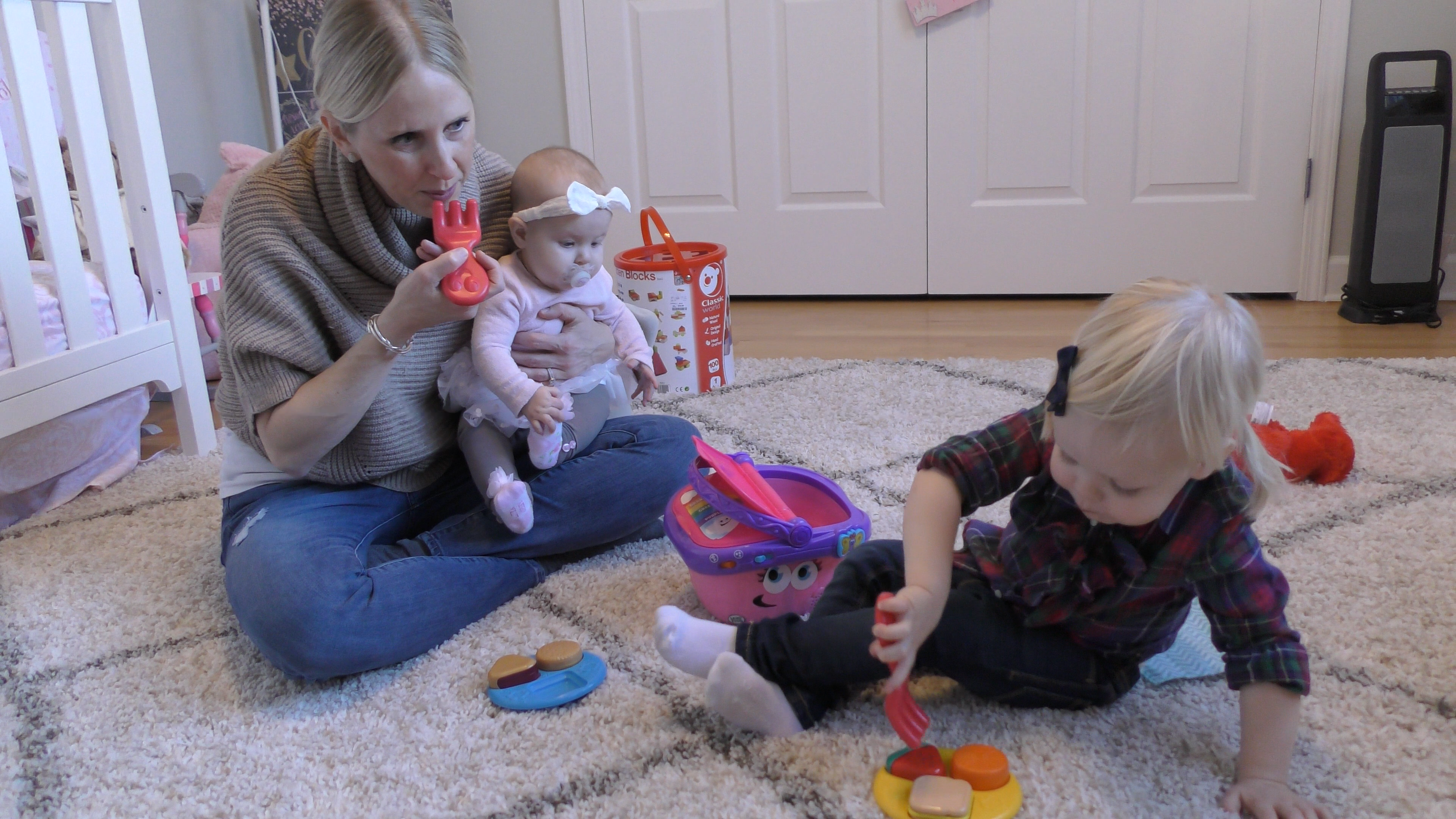 Mom holds baby girl on her lap while playing with pretend food with her toddler daughter on a bedroom floor.