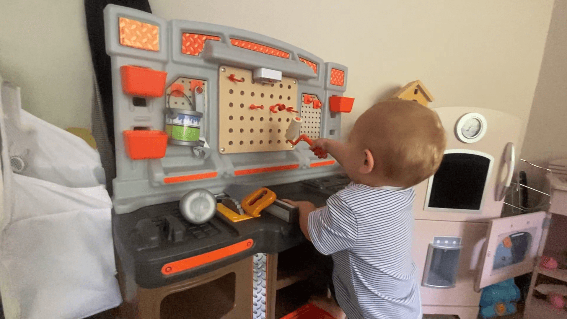 Toddler boy playing with a toy paint roller at a tool bench playset.