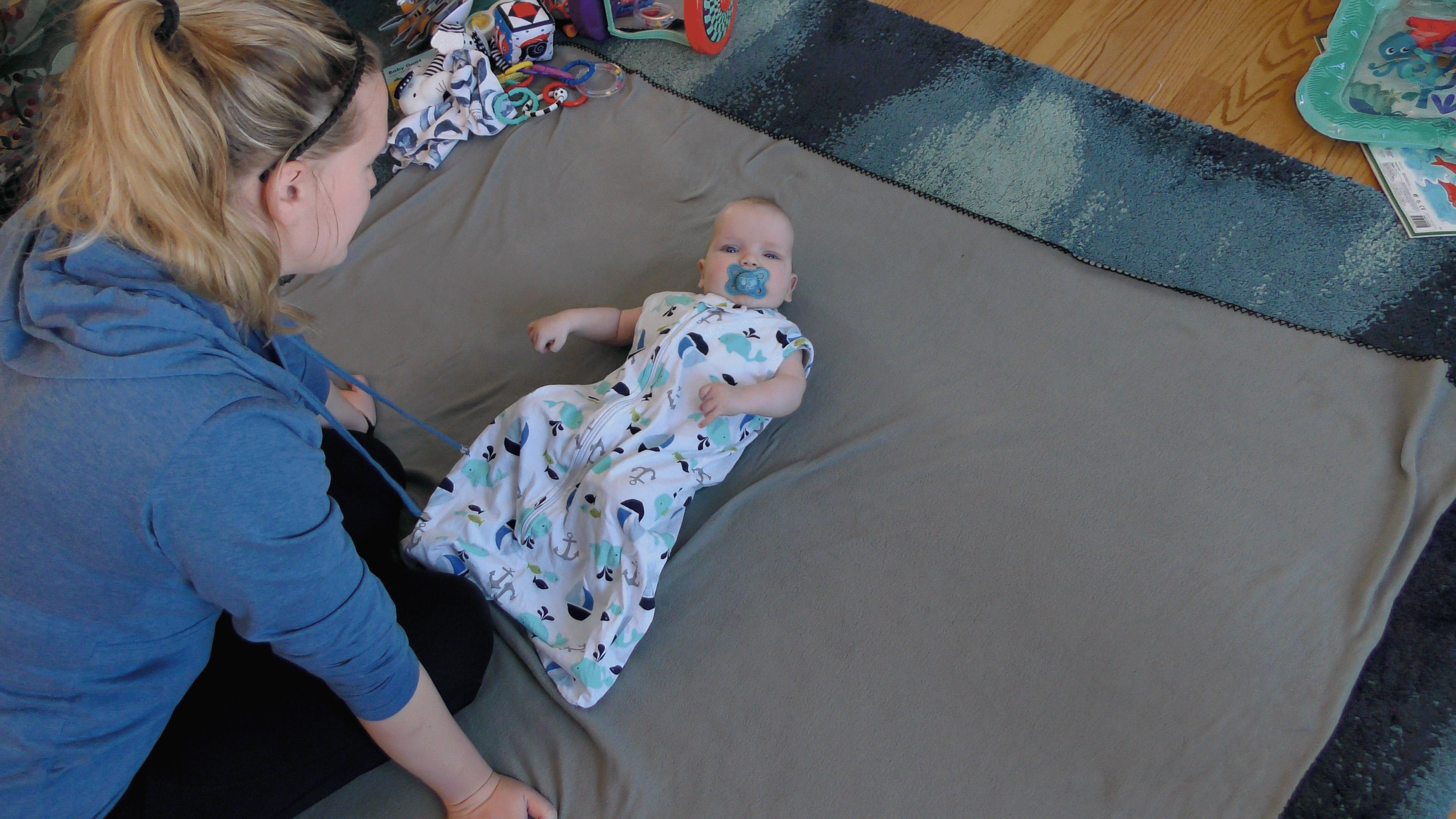 Mom leaning over baby boy dressed in sleep sack on the floor.