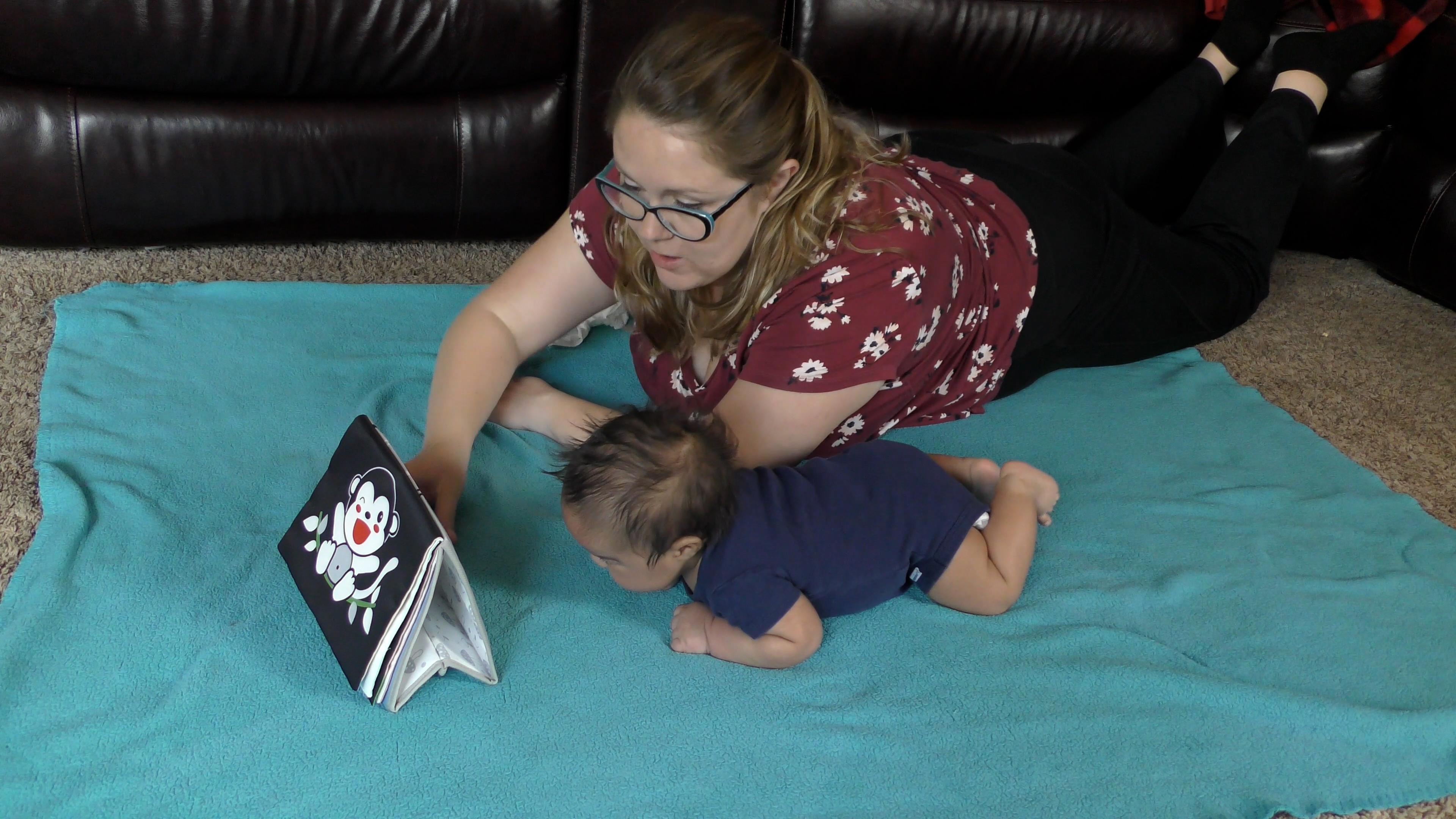 Mom and baby boy lying on tummies together on top of a blue blanket. The mom is pointing at a high-contrast-colored activity book to encourage the baby to lift his head.