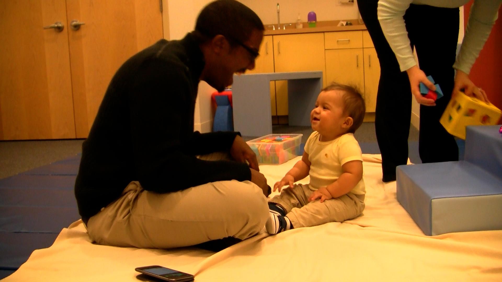Dad and baby boy sitting facing each other and smiling in a play room.