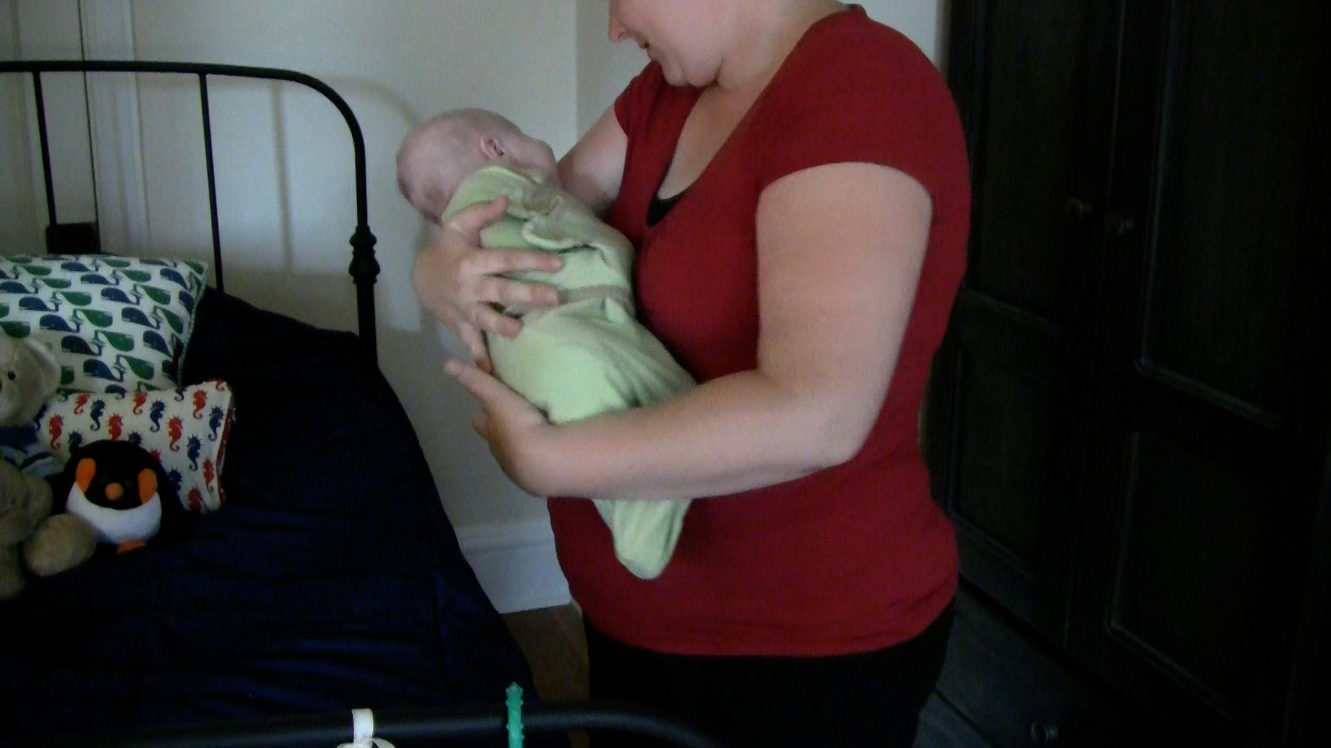 Woman in a red shirt cradles a baby in a light green swaddle. She is standing next to a bed in a dimly lit bedroom.