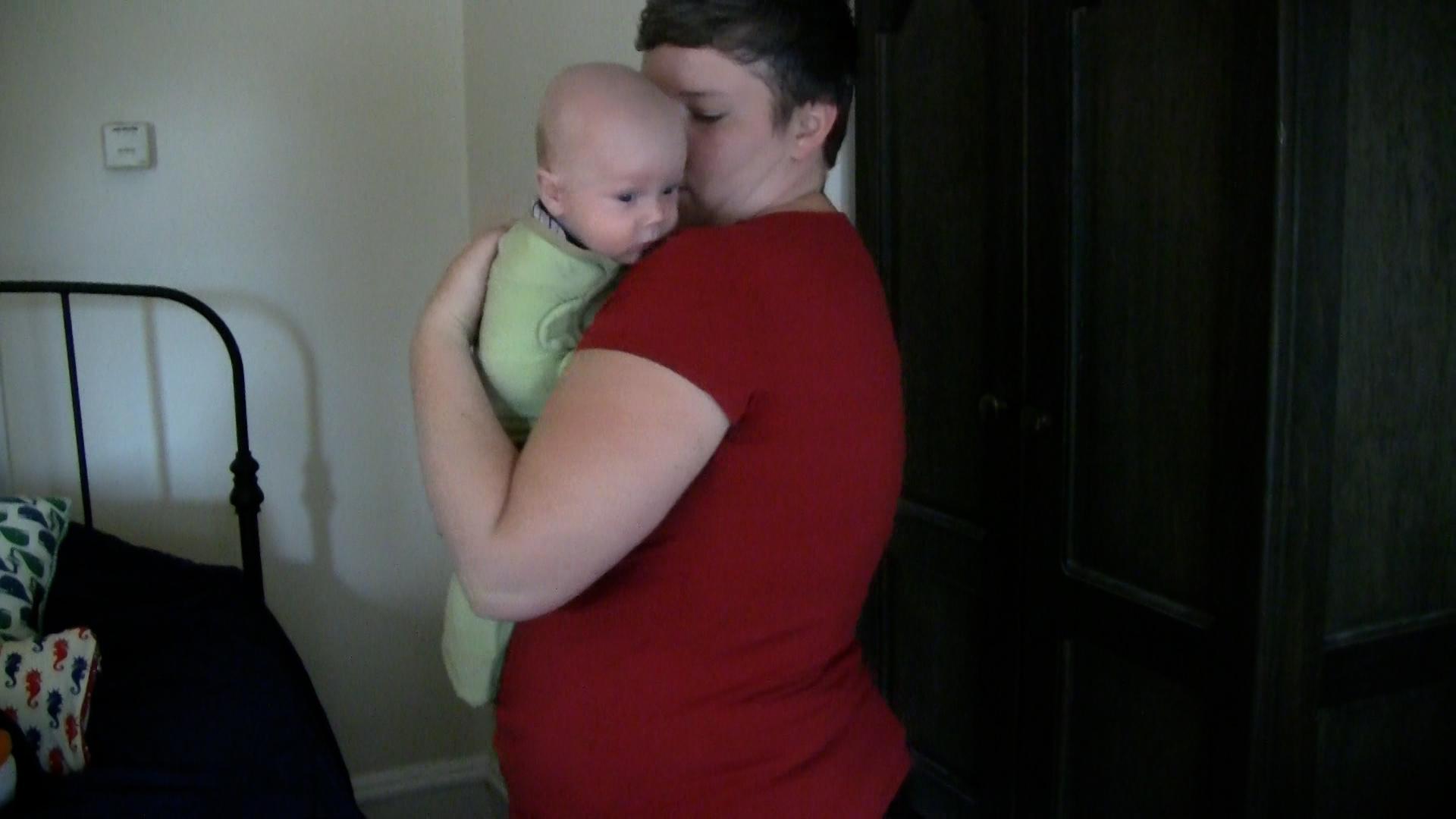 Mom in red t-shirt holding swaddled baby upright over one shoulder.
