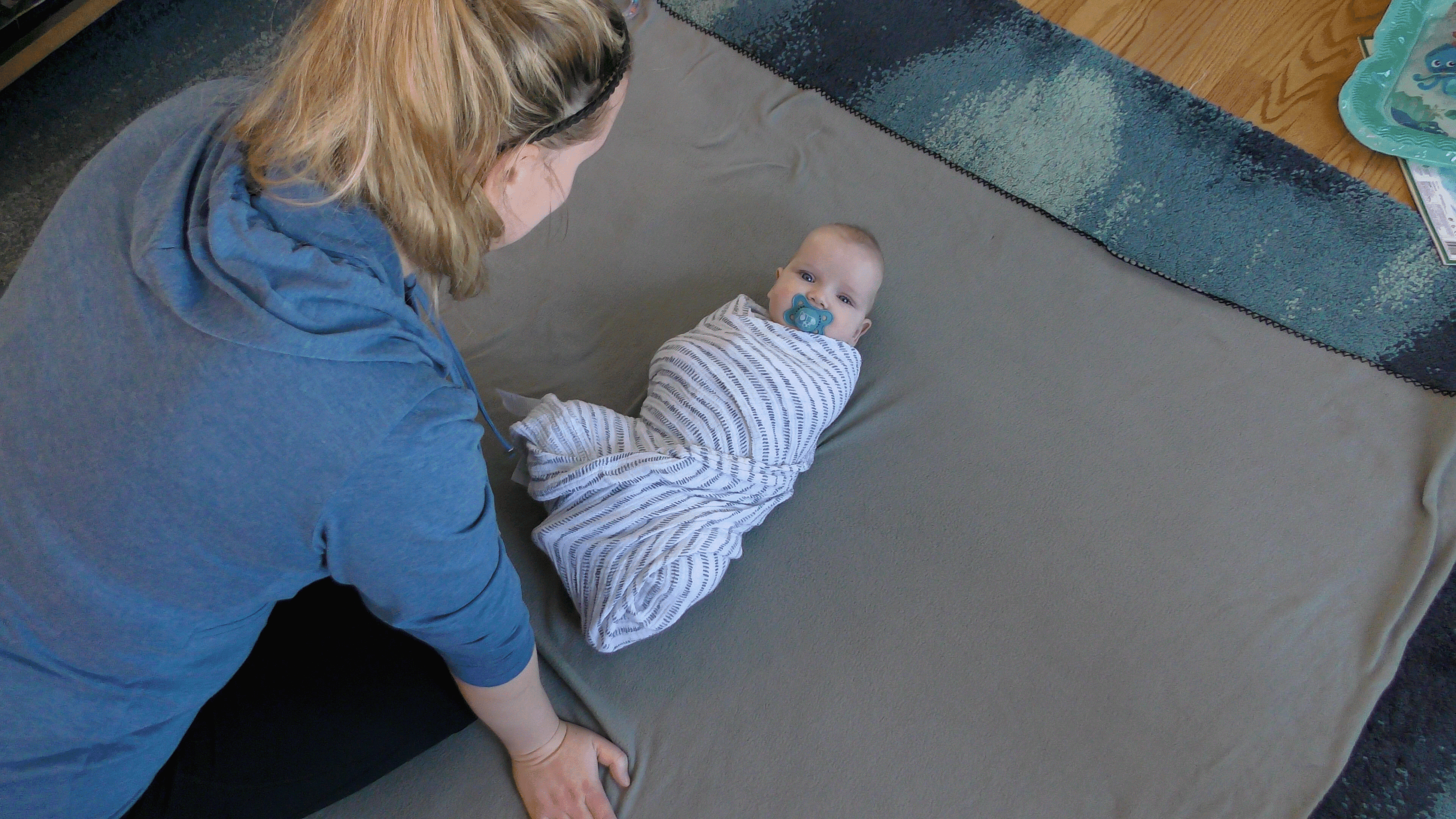 Mom leaning over baby boy wrapped in a swaddle on the floor.