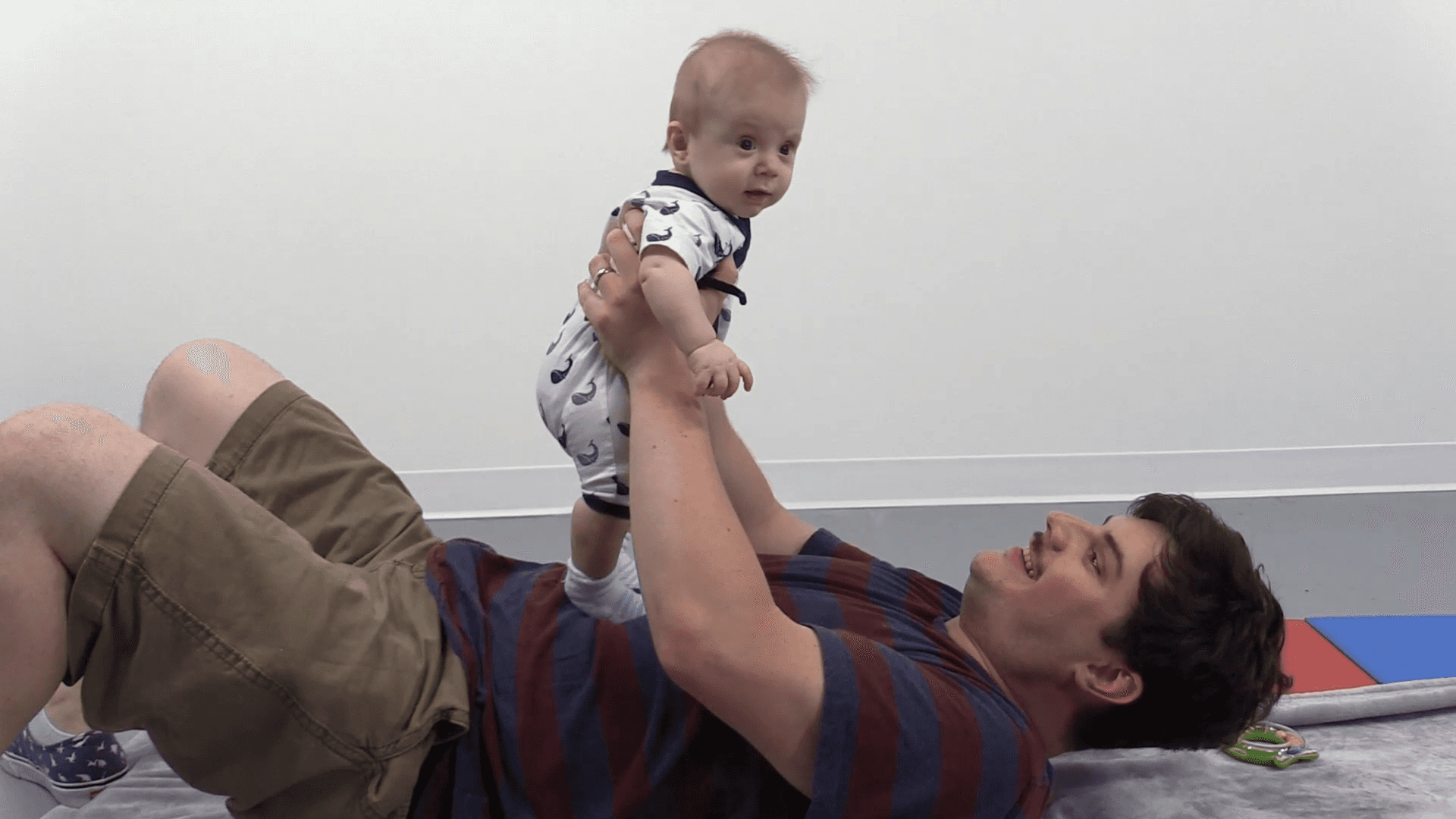 Smiling dad lies on his back while holding a happy baby boy in a standing position on his stomach.