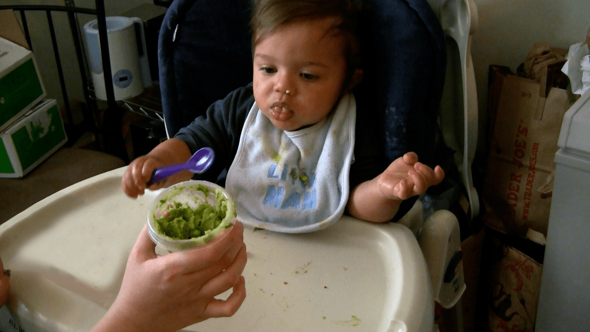Cup of mashed avocado held out to baby boy who is sitting in his high chair with a purple spoon in hand.