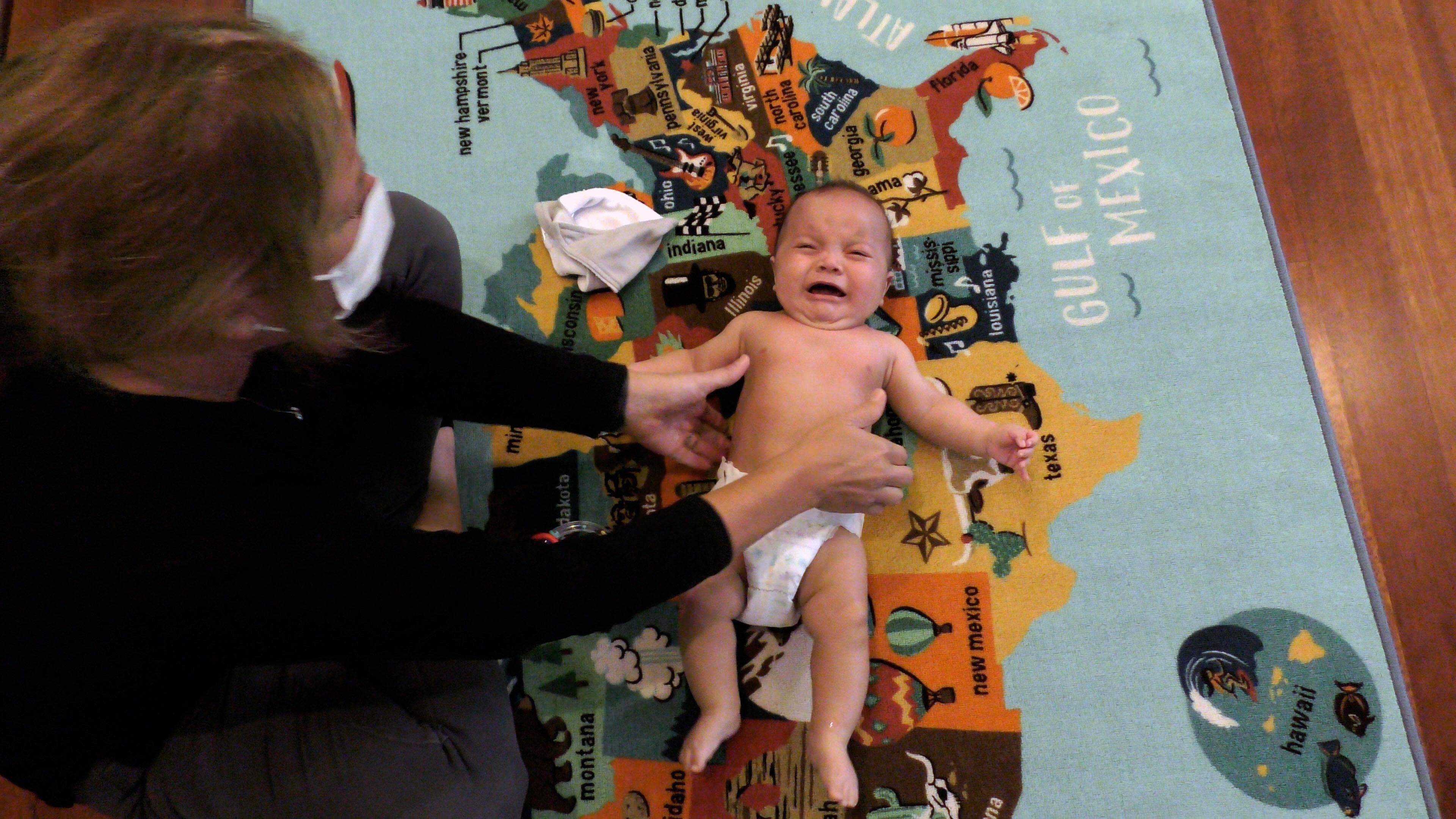 Top-down view of a crying baby boy in a diaper lying on the floor. Beneath him is a colorful kids’ rug showing a map of the United States. A woman sitting cross-legged to the left of the baby reaches her hands out to pick him up.