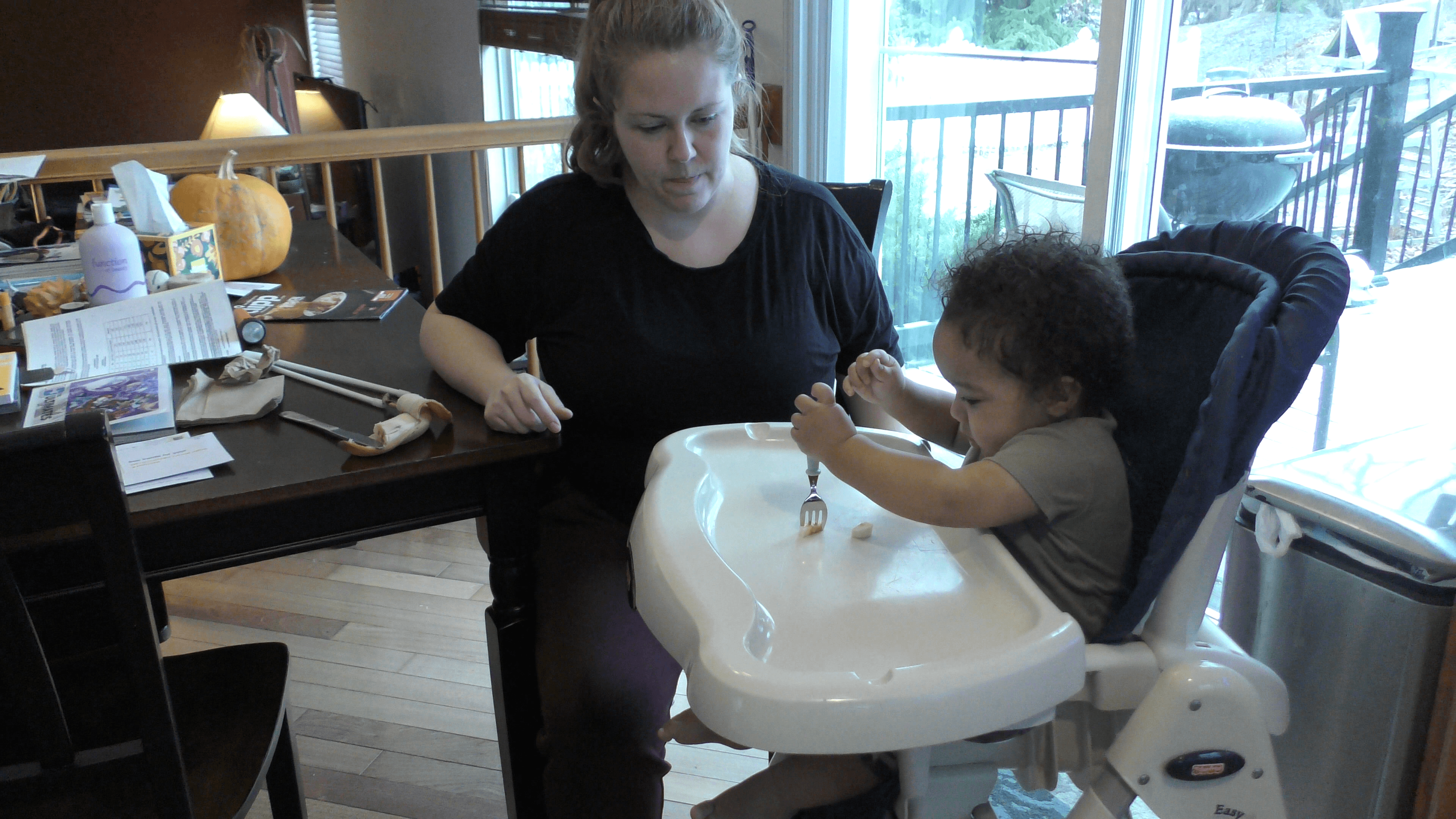 Mom siting at kitchen table watching baby boy attempt to stab a piece of food with a fork on his high chair table.