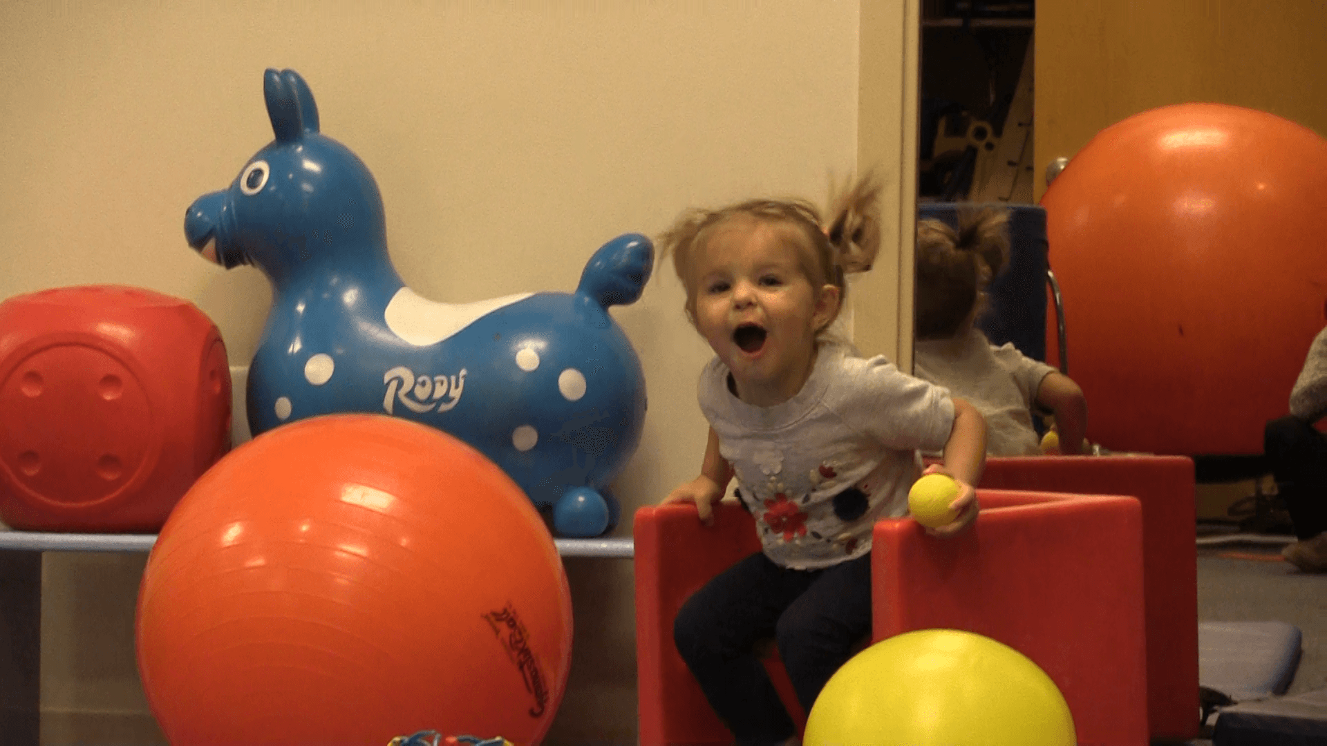 Excited toddler girl playing in a toy room next to balls and toys of different sizes.