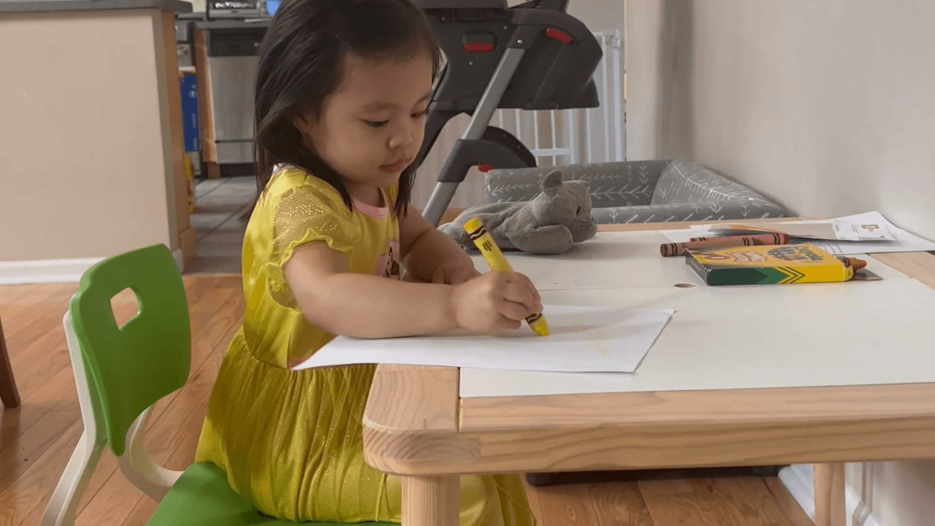 Preschool-aged girl sitting at a child-sized table and drawing with a jumbo yellow crayon.