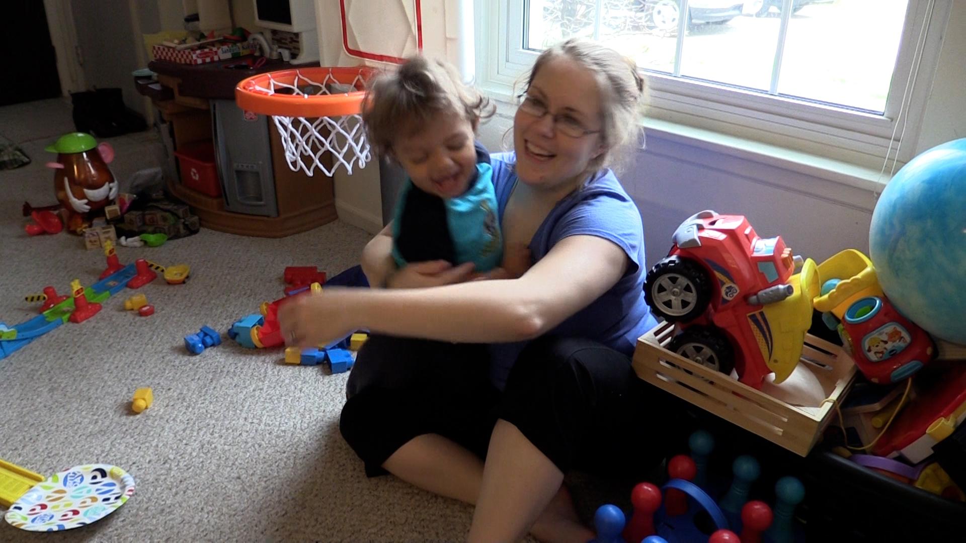 A mom wraps a smiling toddler boy into a big bear hug. The mom and toddler are in a playroom surrounded by toddler toys, including a mini basketball hoop, toy truck, and blocks.