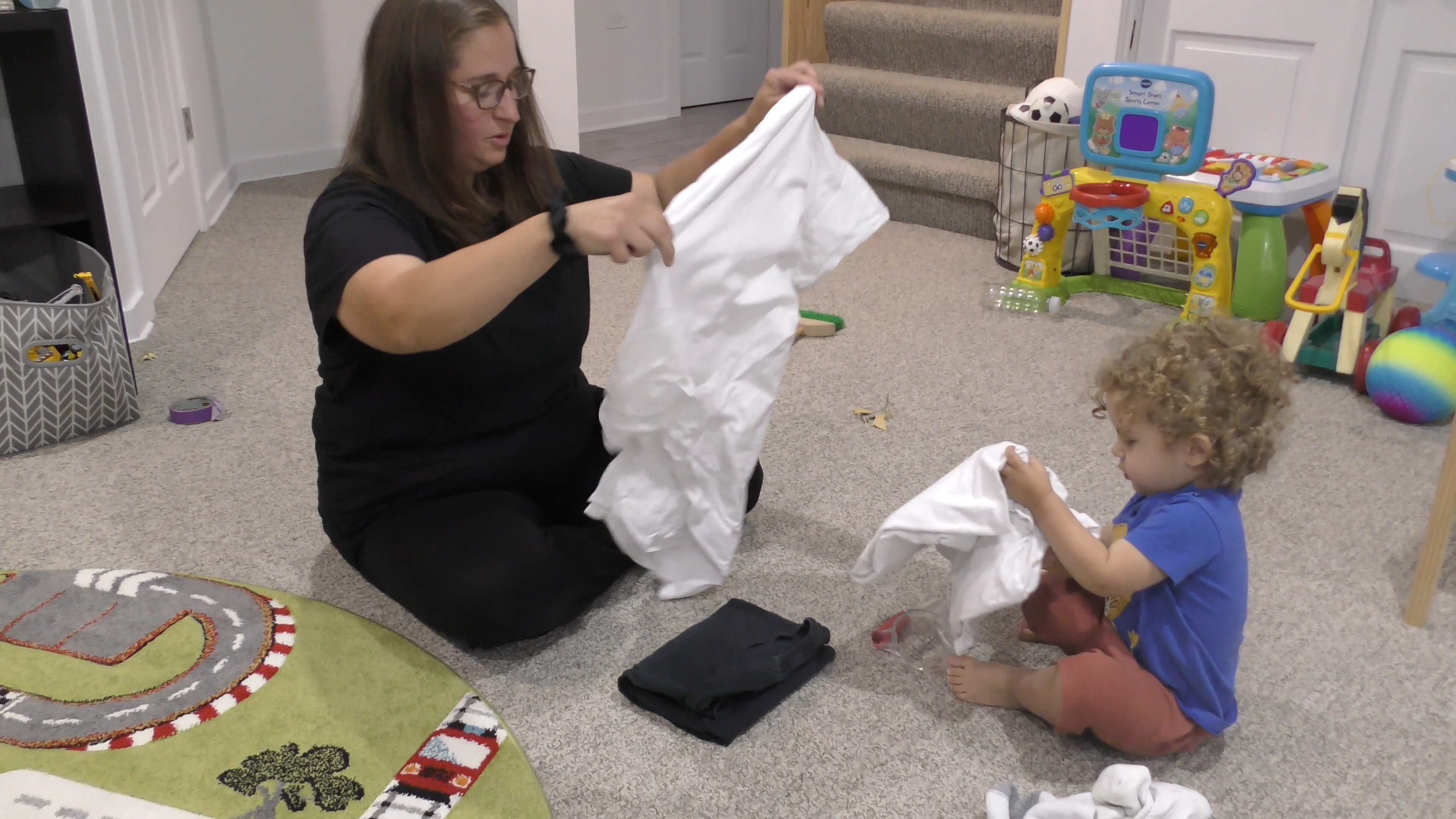 Toddler boy copying mother as she folds laundry on a playroom floor.