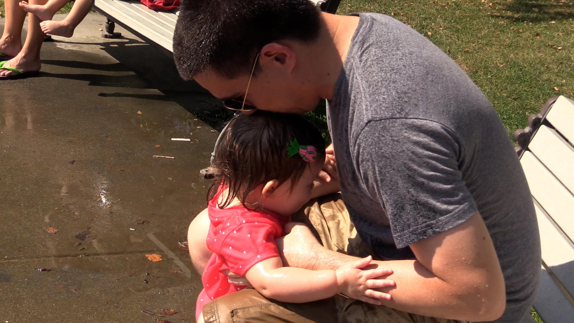 A scared toddler girl clings onto her dad at the park. The dad is sitting on a park bench and kissing the top of his daughter’s head.