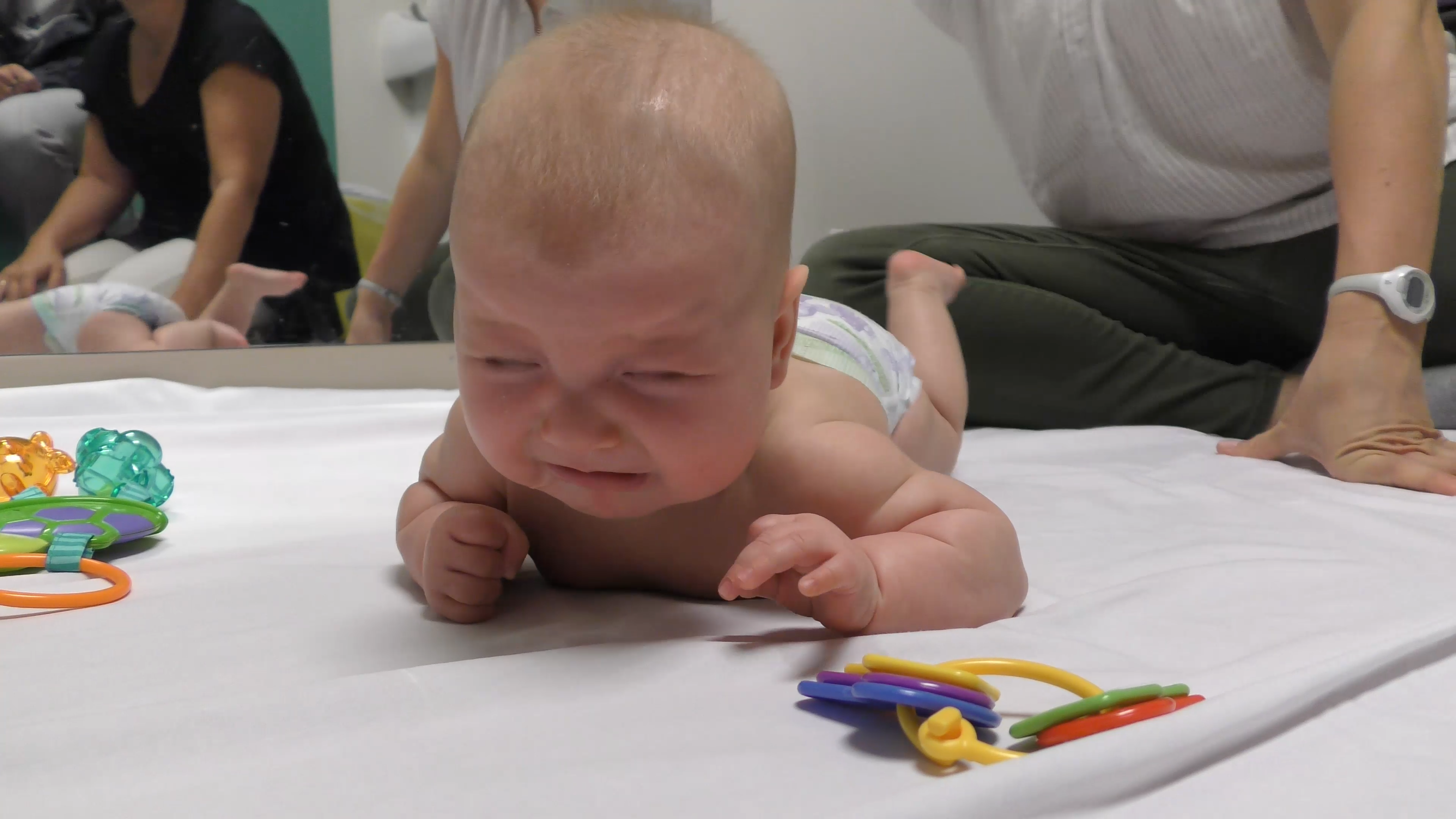 Baby crying while doing Tummy Time. Two caregivers sit nearby.