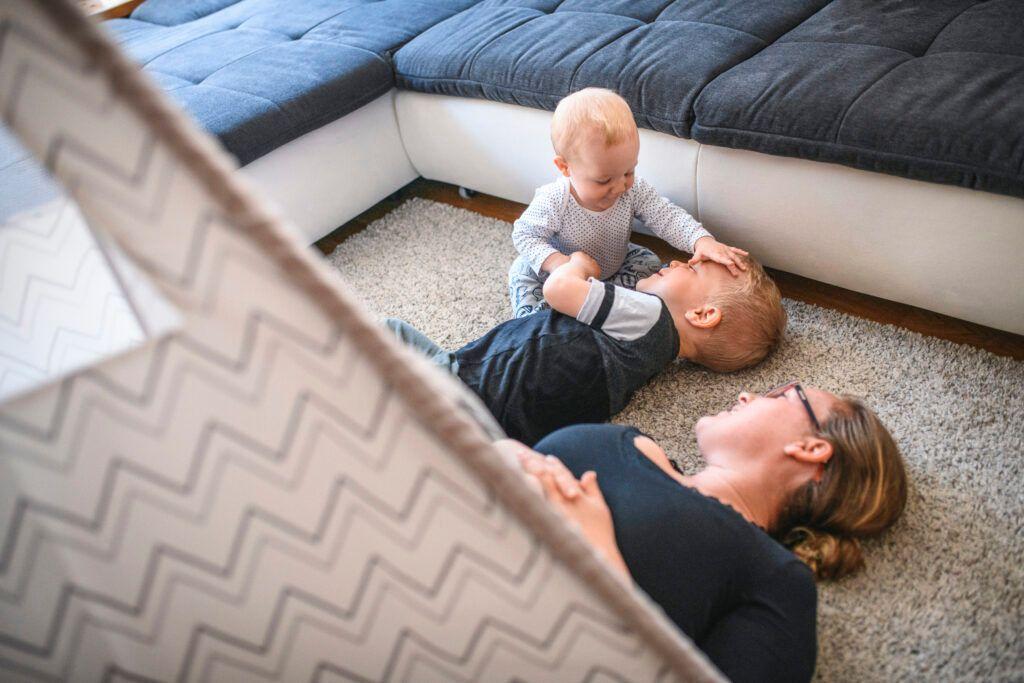 Mom and toddler boy lie next to sitting baby boy who is babbling at his brother.
