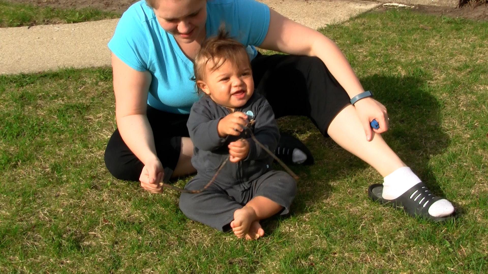 Toddler boy holds up a blue flower while sitting cross-legged in front of his mom on the grass.