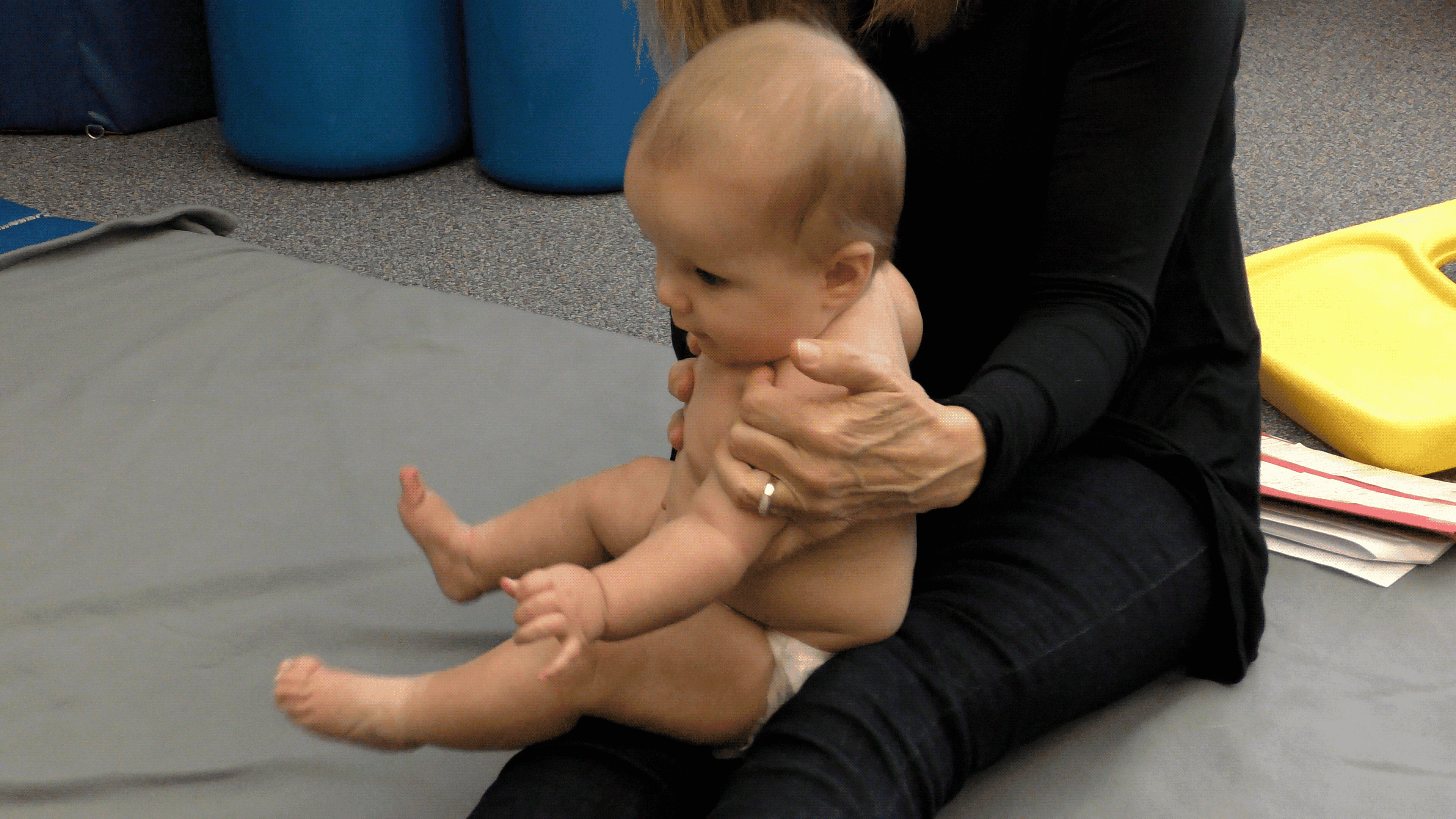 A baby with a flat spot on the back of its head (positional plagiocephaly) is held in a sitting position on a caregiver’s legs.