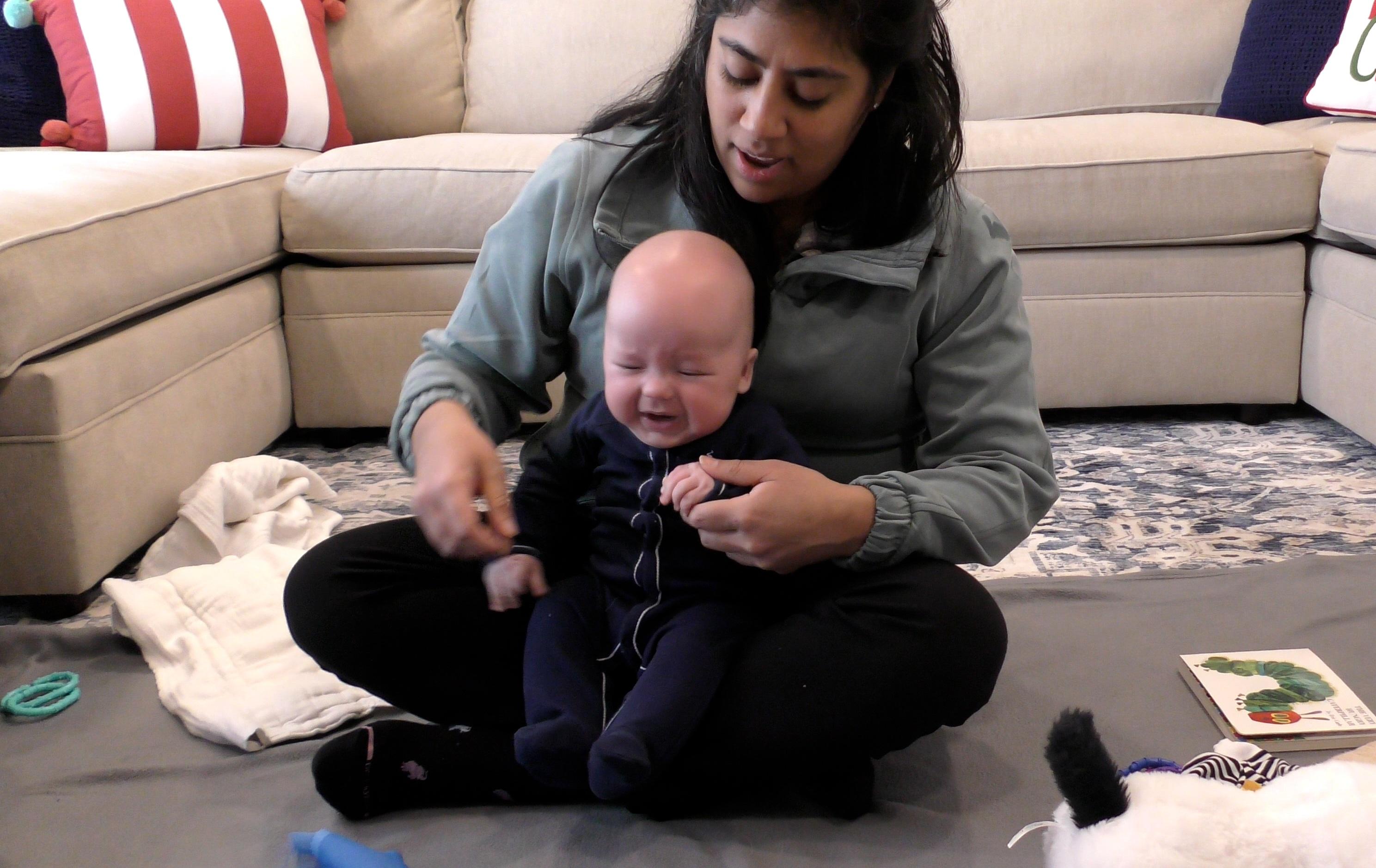 Baby boy sneezing while sitting in a woman’s lap on a living room floor. Toys surround them, and a beige couch fills the background behind them.